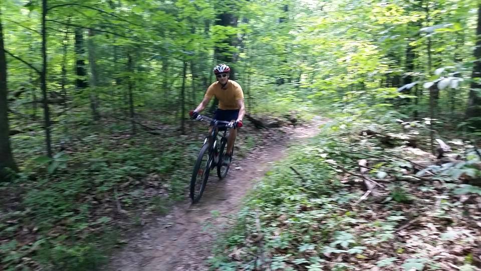 A person riding a mountain bike on a dirt trail through a lush green forest. The cyclist is wearing a yellow shirt and a helmet, navigating the winding path surrounded by trees and foliage. Mohican mountain bike trail.