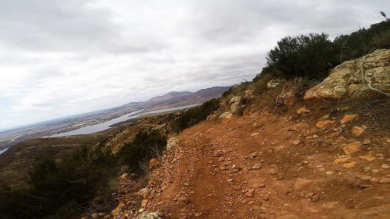 A winding dirt trail bordered by rocky terrain, leading down a hillside with sparse vegetation. In the distance, a large body of water is visible, framed by rolling hills under a cloudy sky. Otay Valley Regional Park mountain bike trail.