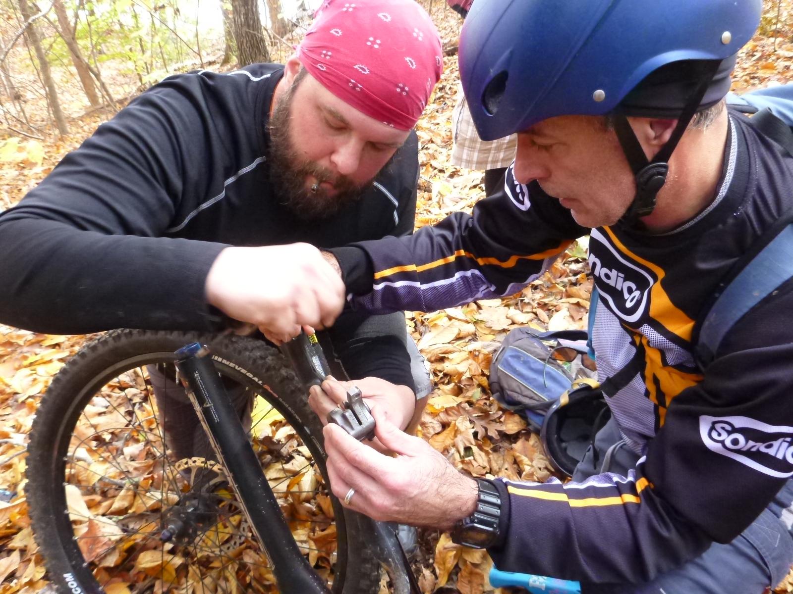 Two individuals are working together to repair a mountain bike in a forested area covered with autumn leaves. One person is focused on adjusting a piece of the bike with a tool, while the other is assisting by holding a component. Both are dressed in casual outdoor attire, and a backpack is visible in the background. Wild Turkey mountain bike trail.