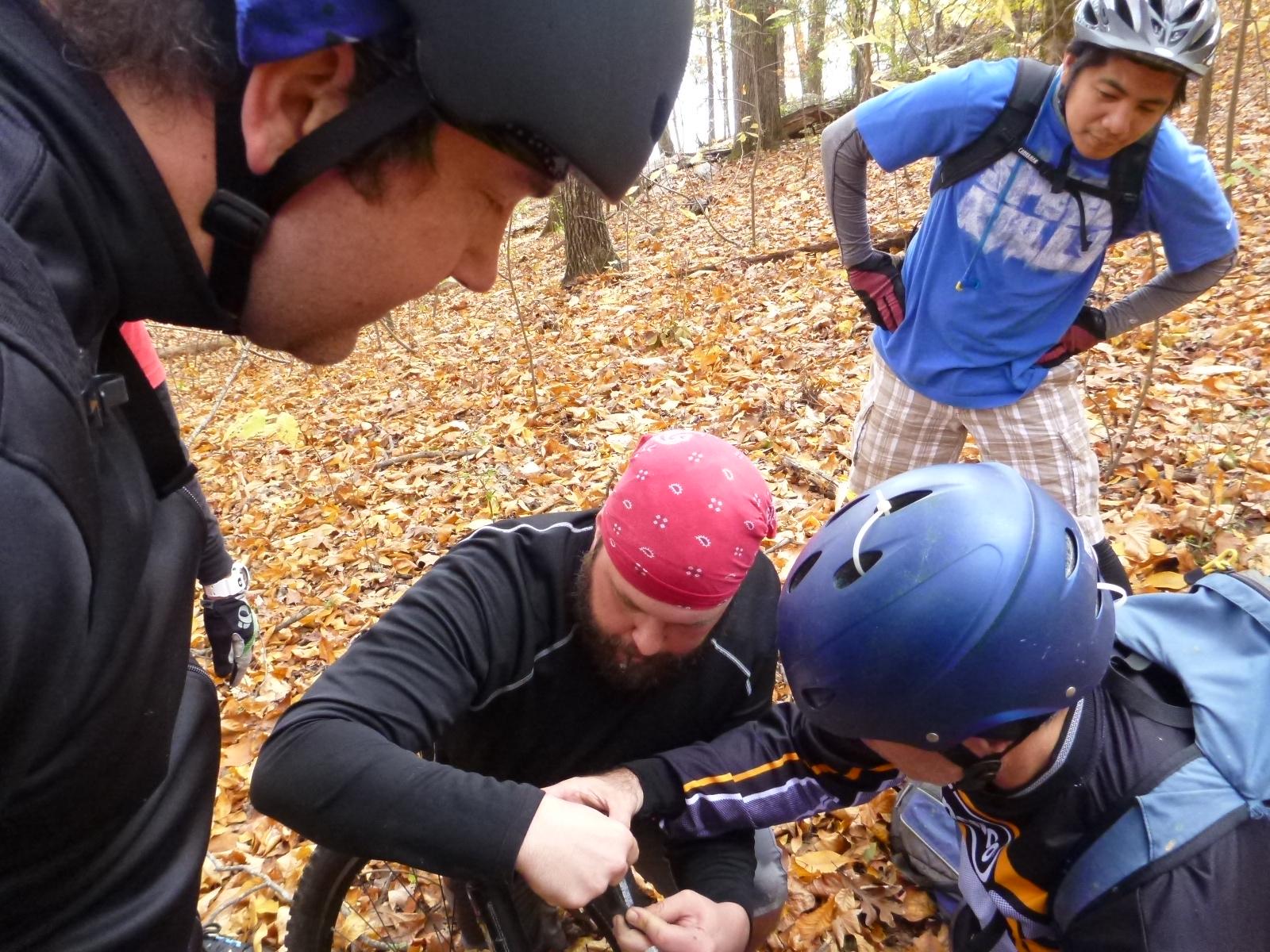 A group of four mountain bikers, wearing helmets and cycling gear, gather around a bike on a trail covered in autumn leaves. One person, in a red bandana, is crouched down working on the bike, while the others watch attentively. The scene is set in a forested area with fall foliage in the background. Wild Turkey mountain bike trail.