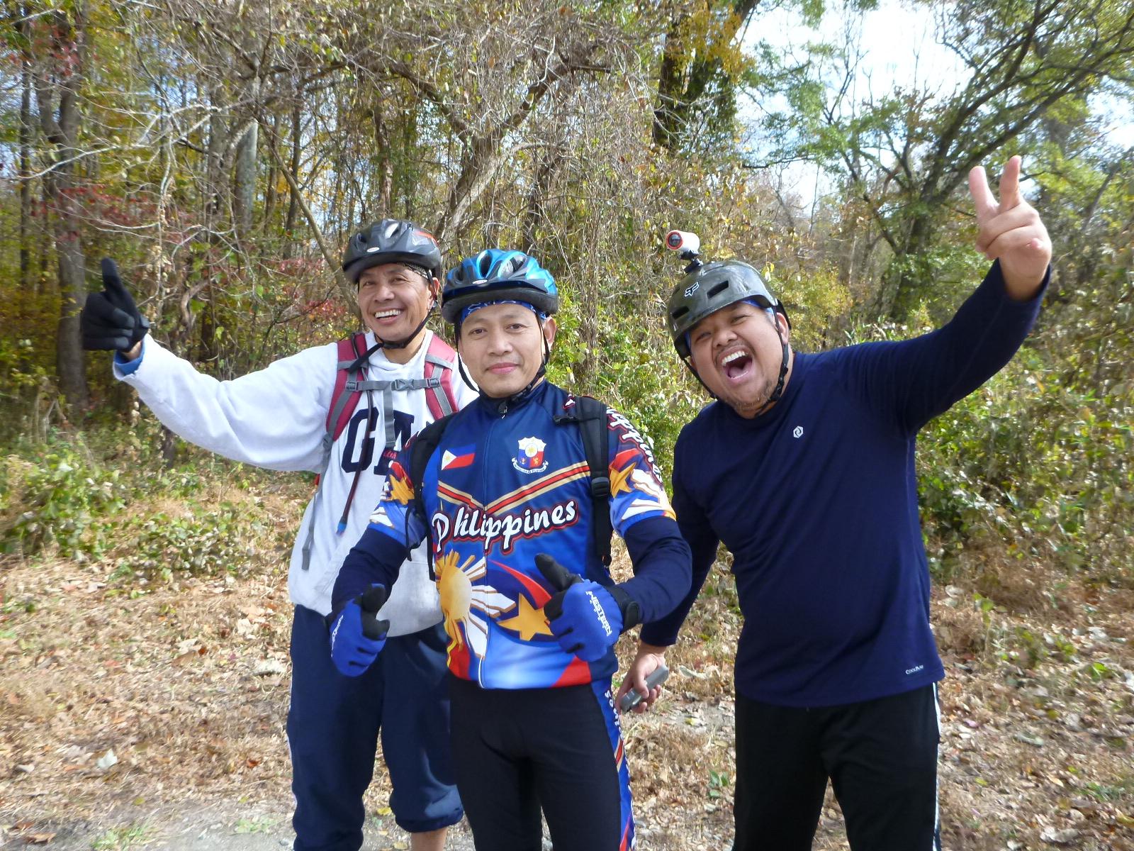 Three men in cycling gear pose for a photo outdoors, surrounded by trees. One man is wearing a helmet and a brightly colored jersey with the word "Philippines," while the others are dressed casually, with one giving a thumbs-up and another making a peace sign. The group displays cheerful expressions, suggesting a fun and active day outdoors. Wild Turkey mountain bike trail.