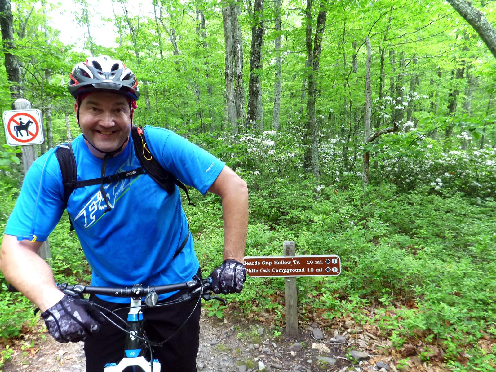 A smiling person in a blue cycling jersey and helmet is posing with a mountain bike on a trail surrounded by lush green trees. In the background, there's a sign indicating trail directions and a no horses allowed sign. The scene is vibrant and represents an outdoor biking adventure. Douthat State Park mountain bike trail.