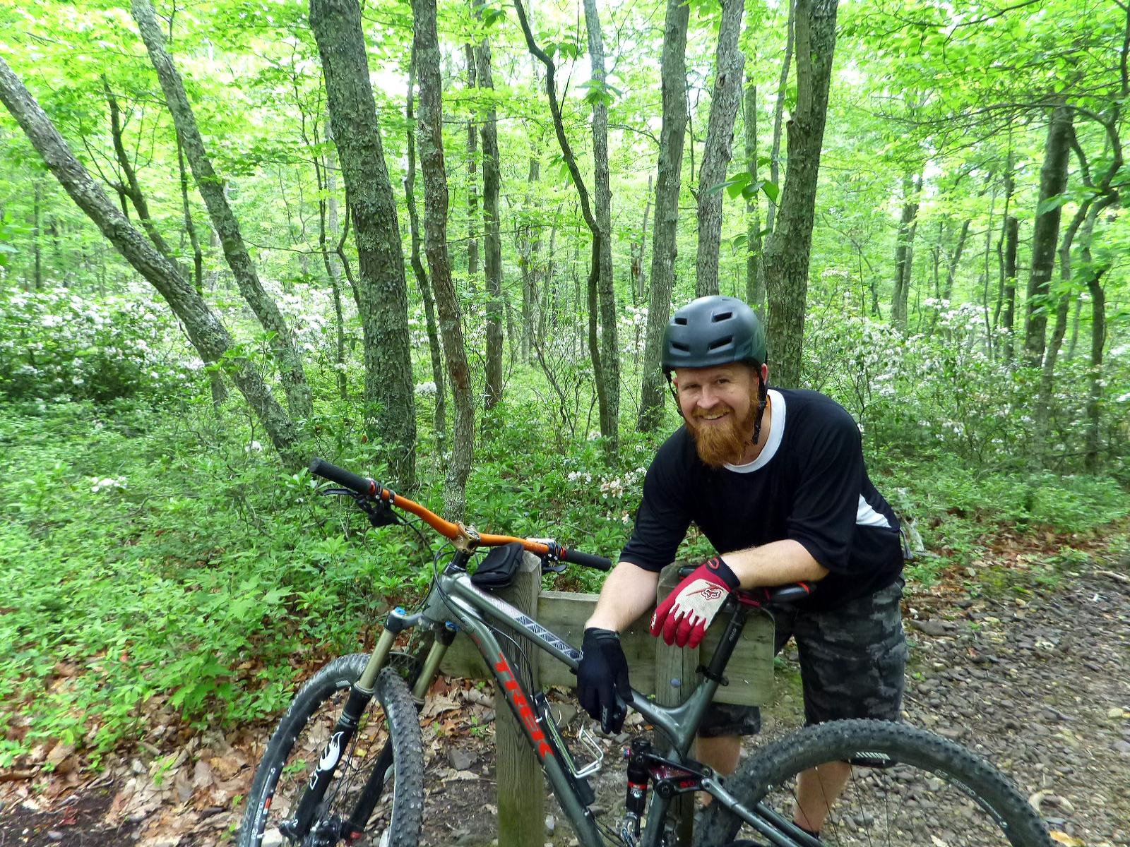 A man with a beard and a helmet smiles while leaning against his mountain bike in a lush, green forest. The background features trees and blooming flowers, creating a vibrant outdoor scene. He is wearing a black shirt, camouflage shorts, and gloves, presenting a relaxed and enthusiastic demeanor. Douthat State Park mountain bike trail.