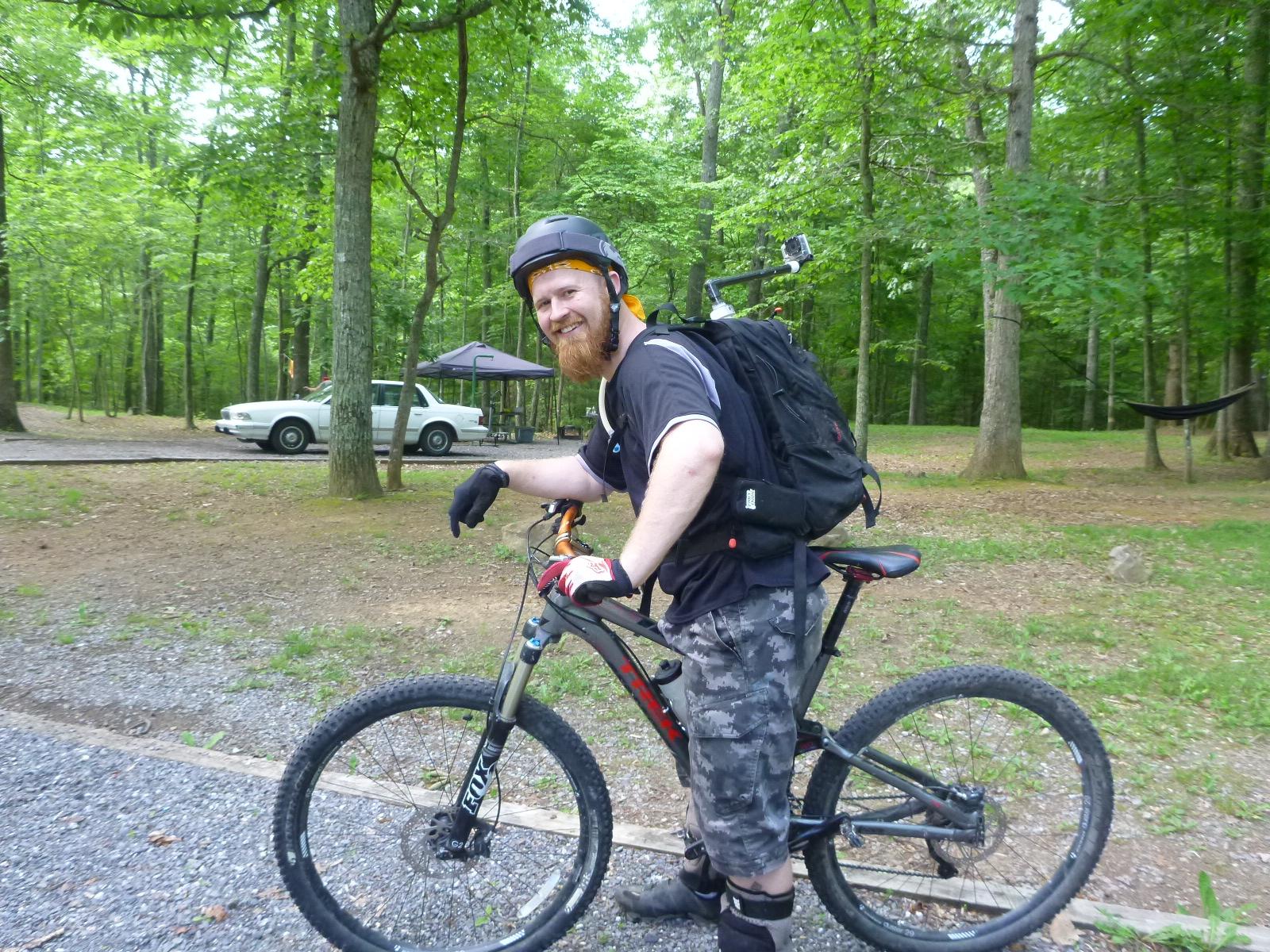 An individual with a red beard wearing a helmet and gloves is standing beside a mountain bike in a forested area. They are smiling and wearing casual clothing, including a black shirt and camouflage shorts. In the background, there are trees, a parked white car, and a hammock. Douthat State Park mountain bike trail.