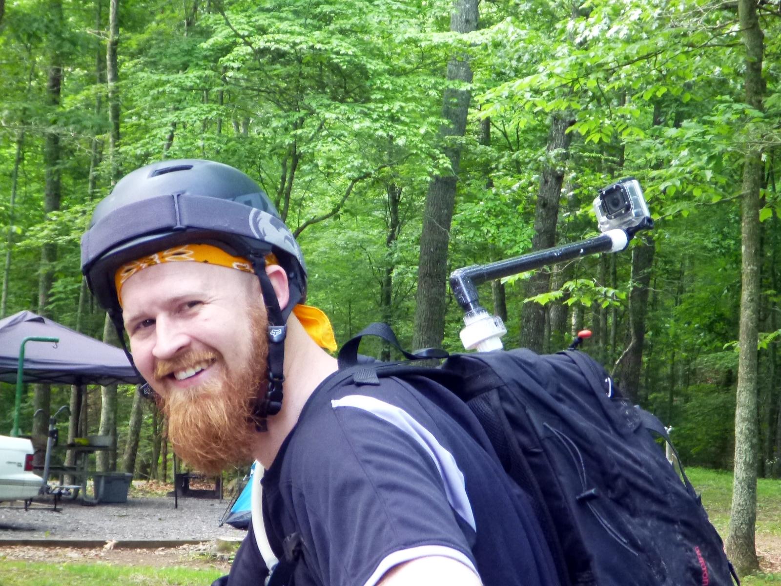 A smiling man with a beard wearing a helmet and a bandana stands outdoors in a forested area. He has a camera mounted on a pole attached to his backpack. Green trees and camping equipment are visible in the background. Douthat State Park mountain bike trail.