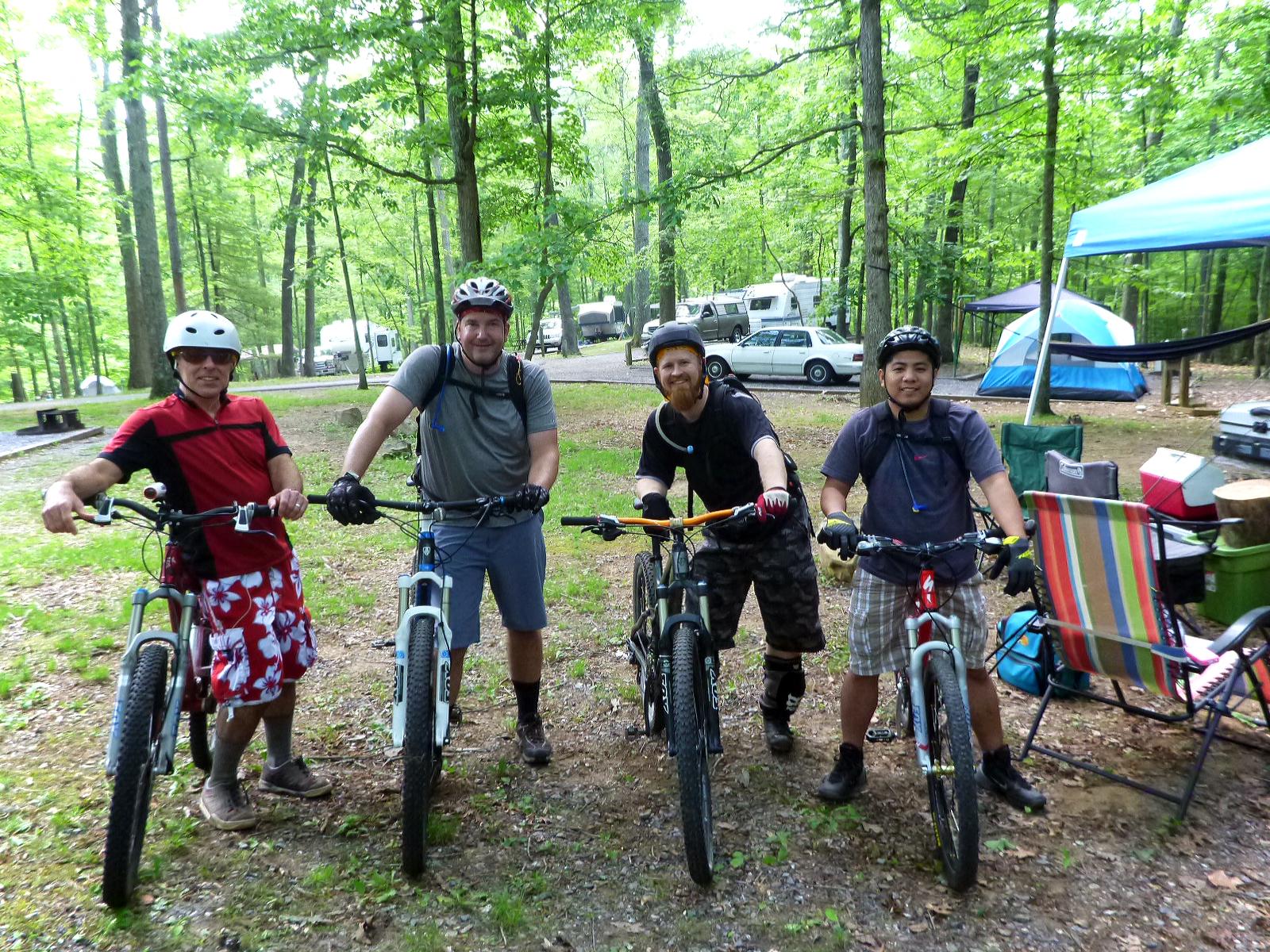 Four men stand with mountain bikes in a wooded camping area. They are dressed in casual outdoor clothing and helmets, smiling at the camera. In the background, there are tents and vehicles visible among the trees, suggesting a recreational atmosphere. The ground is covered with grass and dirt, and it appears to be a sunny day. Douthat State Park mountain bike trail.