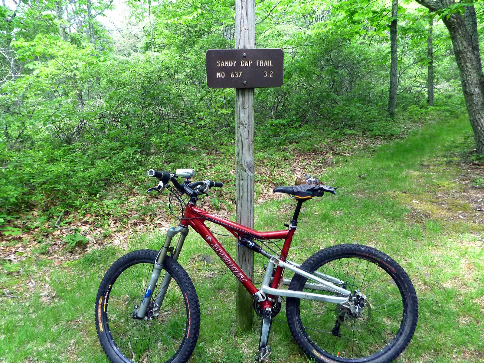 Mountain bike resting beside a trail sign for Sandy Gap Trail, which indicates a distance of 3.2 miles. The surrounding area features lush green vegetation and trees. Douthat State Park mountain bike trail.