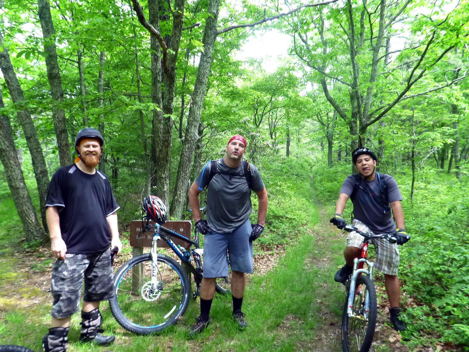 Three men take a break from mountain biking on a forested trail. They are wearing helmets and biking gear, standing next to their bikes with trees and greenery surrounding them. One man is smiling, while the others look relaxed. A trail sign is visible in the background. Douthat State Park mountain bike trail.