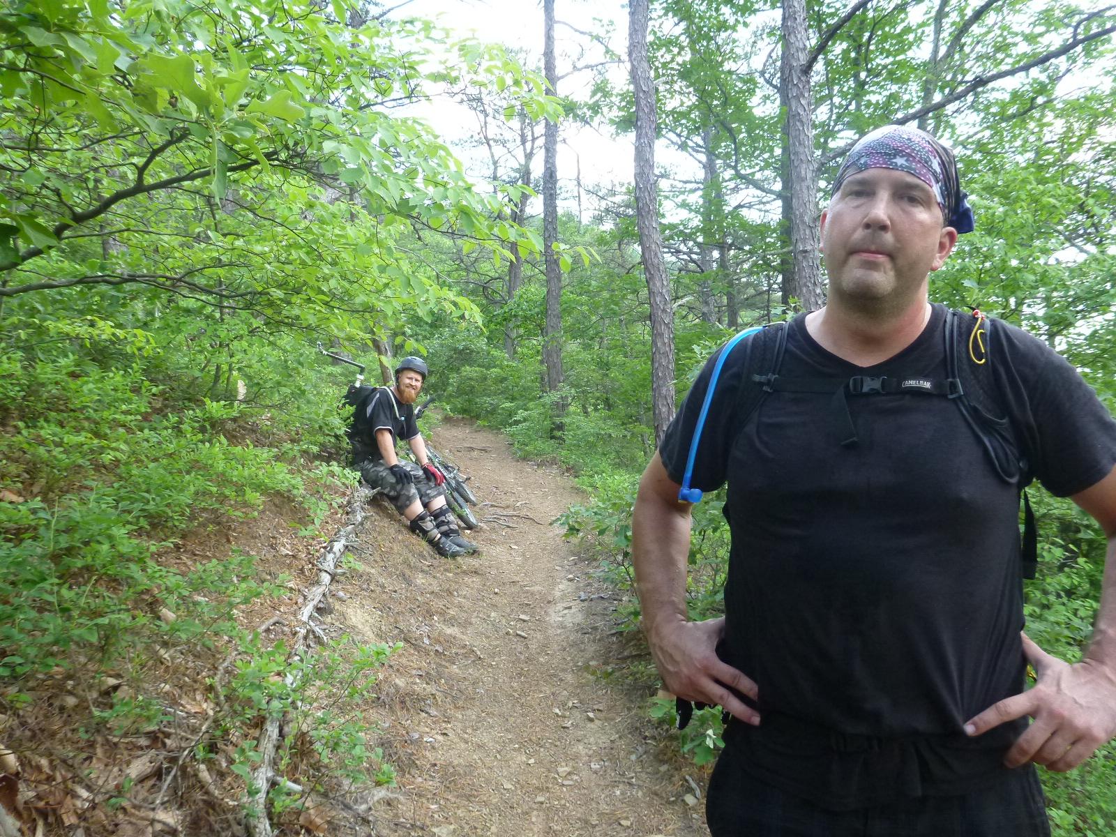 Two individuals are resting on a hiking trail surrounded by lush green foliage. One person is sitting on the ground with a bike beside him, wearing a helmet and casual outdoor clothing. The other person is standing nearby, dressed in a black shirt and carrying a backpack with water tubing. The setting features a mix of trees and underbrush, indicating a mountainous or forested area. Douthat State Park mountain bike trail.
