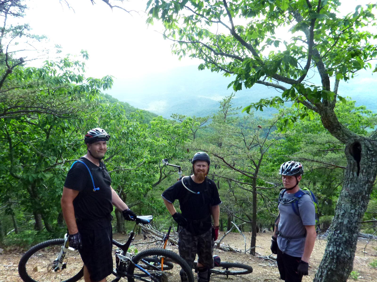 Three mountain bikers pause for a photo while resting on a trail overlooking a lush green valley. They are wearing helmets and biking gear, with their bikes parked nearby. The background features trees and a misty landscape, giving a sense of adventure and outdoor activity. Douthat State Park mountain bike trail.