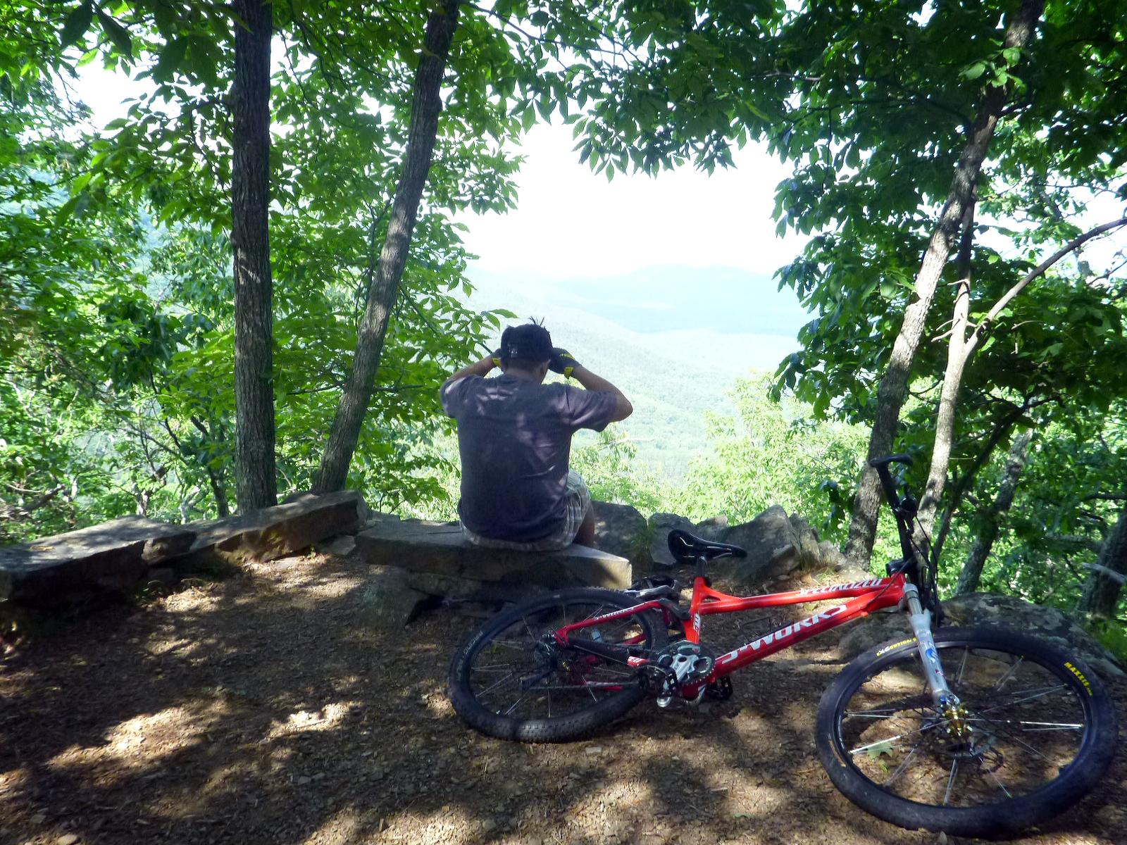 A person sitting on a stone bench in a forest, taking in a scenic view of rolling hills and trees. A red mountain bike is resting on the ground nearby, surrounded by a natural setting of green foliage and sunlight filtering through the leaves. Douthat State Park mountain bike trail.