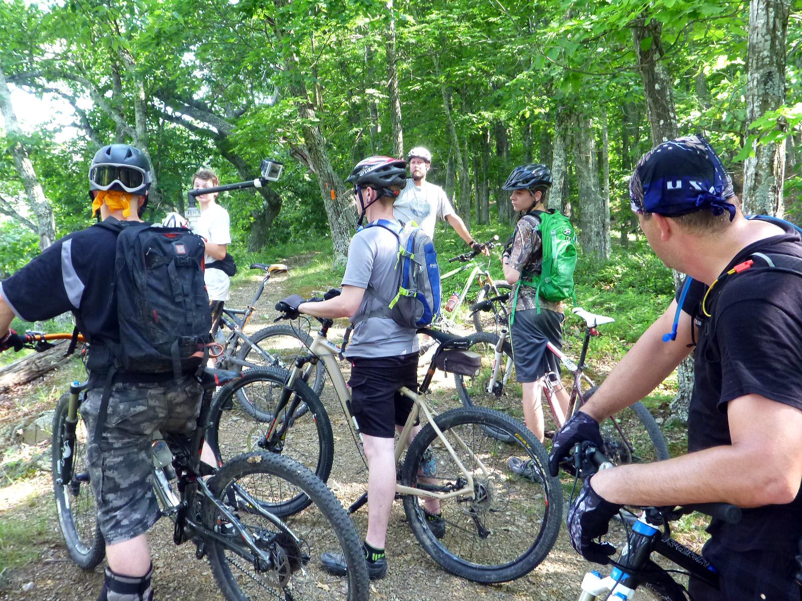 A group of six mountain bikers gathered on a dirt trail in a lush green forest. They are positioned with their bikes, wearing helmets and backpacks. One rider is adjusting a camera mounted on a pole, while others are engaged in conversation, ready to continue their ride. Sunlight filters through the trees, creating a vibrant outdoor scene. Douthat State Park mountain bike trail.