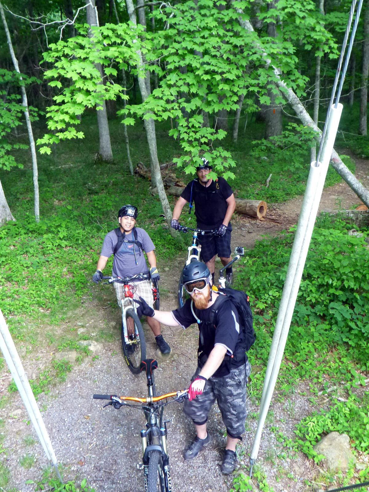 Three mountain bikers in a lush green forest, preparing for a ride. Two riders are standing next to their bikes, with one adjusting his bike's handlebar. They are wearing helmets and safety gear. The background features trees with vibrant green leaves and a gravel path winding through the woods. Douthat State Park mountain bike trail.