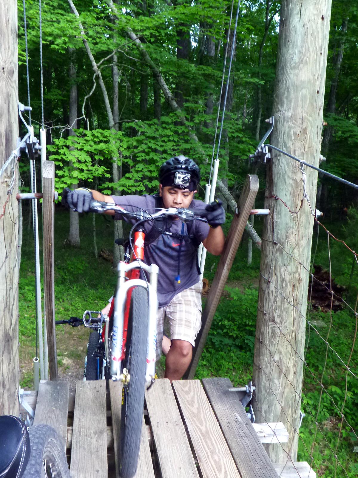 A mountain biker wearing a helmet and gloves navigates a wooden bridge in a forested area. The biker is focused on lifting their bike onto the platform, surrounded by lush green trees. Douthat State Park mountain bike trail.