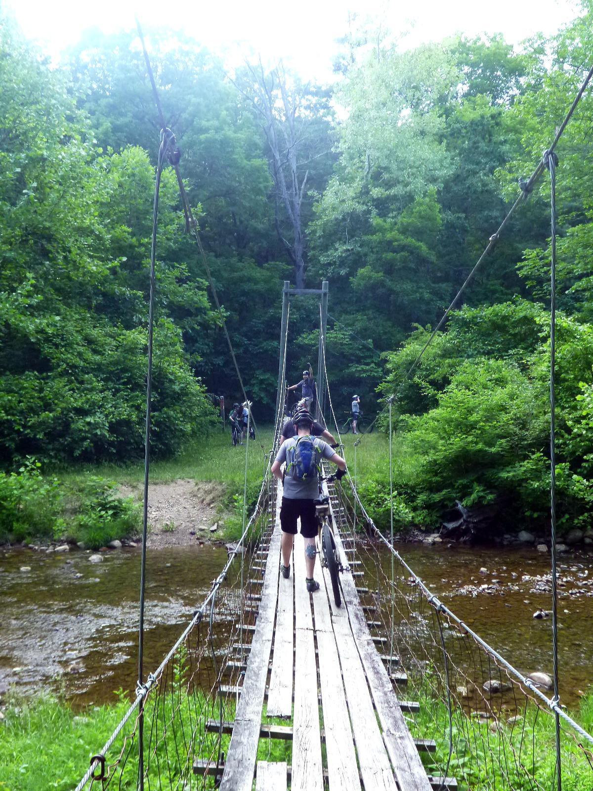 A group of mountain bikers crossing a wooden suspension bridge over a shallow stream, surrounded by lush green trees and foliage. Douthat State Park mountain bike trail.