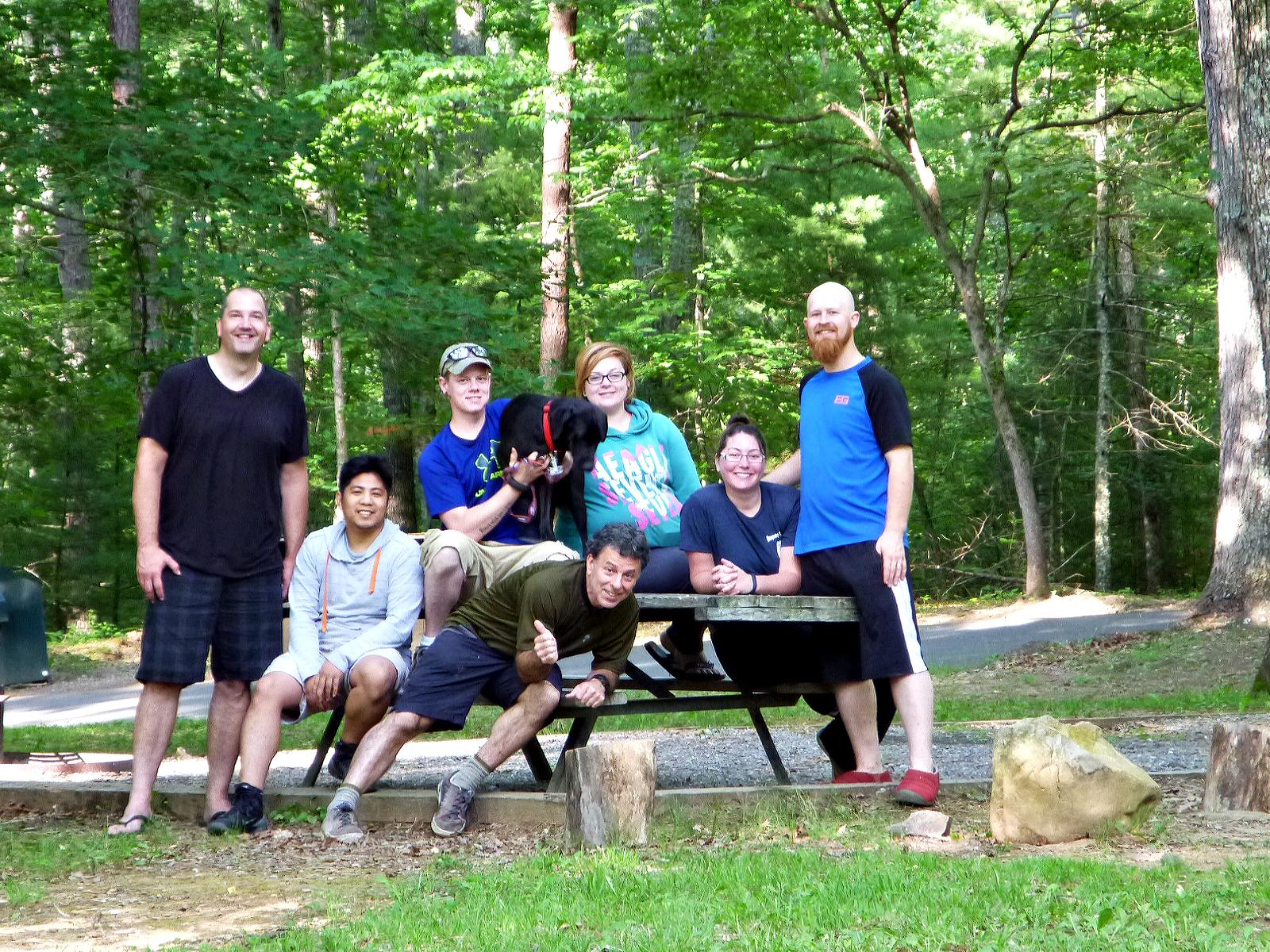 A group of seven people and a dog posing together at a picnic table in a wooded area. The setting features lush green trees and a pathway in the background. The group includes individuals of varying ages, with some sitting and others standing, all smiling and enjoying the moment. Douthat State Park mountain bike trail.