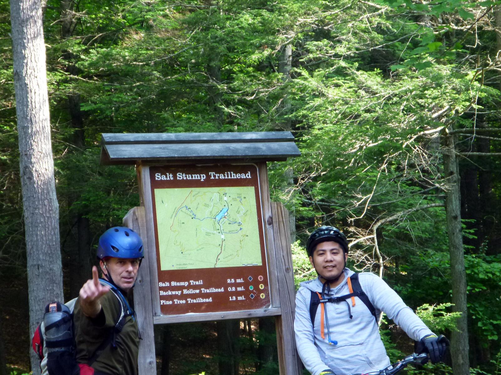 Two mountain bikers pose at the Salt Stump Trailhead sign in a wooded area. One biker, wearing a helmet and casual outdoor attire, points to the sign, while the other, also in a helmet, stands beside a bike. The sign displays a map and details about the trails, surrounded by lush green trees. Douthat State Park mountain bike trail.