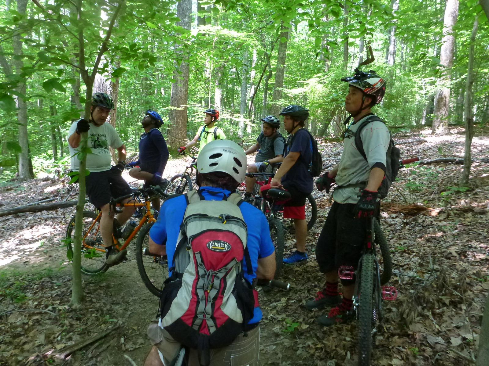 A group of cyclists paused on a wooded trail, surrounded by lush green foliage. They are wearing helmets and biking gear, with some standing beside their mountain bikes. One cyclist is gesturing as if explaining something to the group, while others listen intently. The scene conveys a sense of camaraderie and outdoor adventure. Kernersville MTB park mountain bike trail.