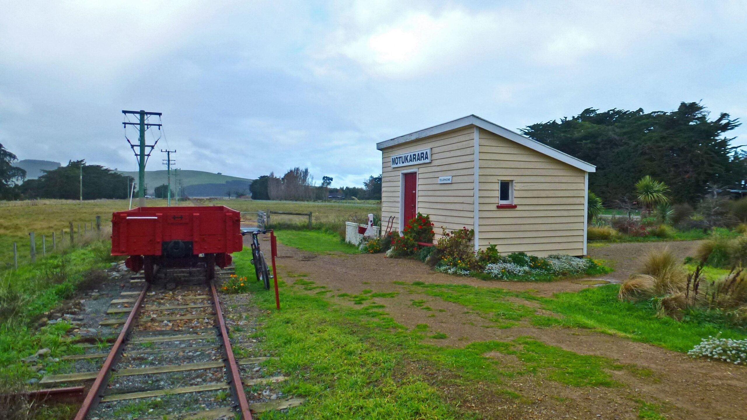 Image of a small, quaint train station building labeled "MOTUKARARA," set alongside railway tracks. A red train car is positioned on the tracks, with grassy fields and rolling hills in the background. Power poles line the path, and the area is adorned with flowers and low shrubs. The sky is overcast, adding a serene atmosphere to the scene. Little River Rail Trail mountain bike trail.