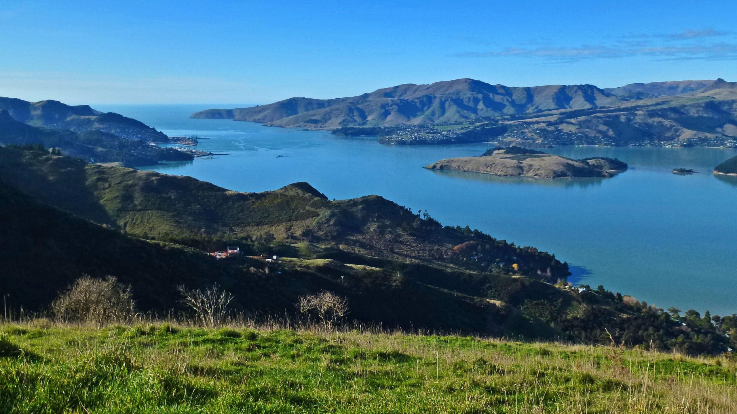 A panoramic view of a tranquil bay surrounded by rolling hills and mountains, with clear blue water and a few small islands visible. The sky is bright with minimal clouds, and a small town can be seen nestled along the shoreline. Lush greenery in the foreground adds to the natural beauty of the scene. Port Hills Tracks mountain bike trail.