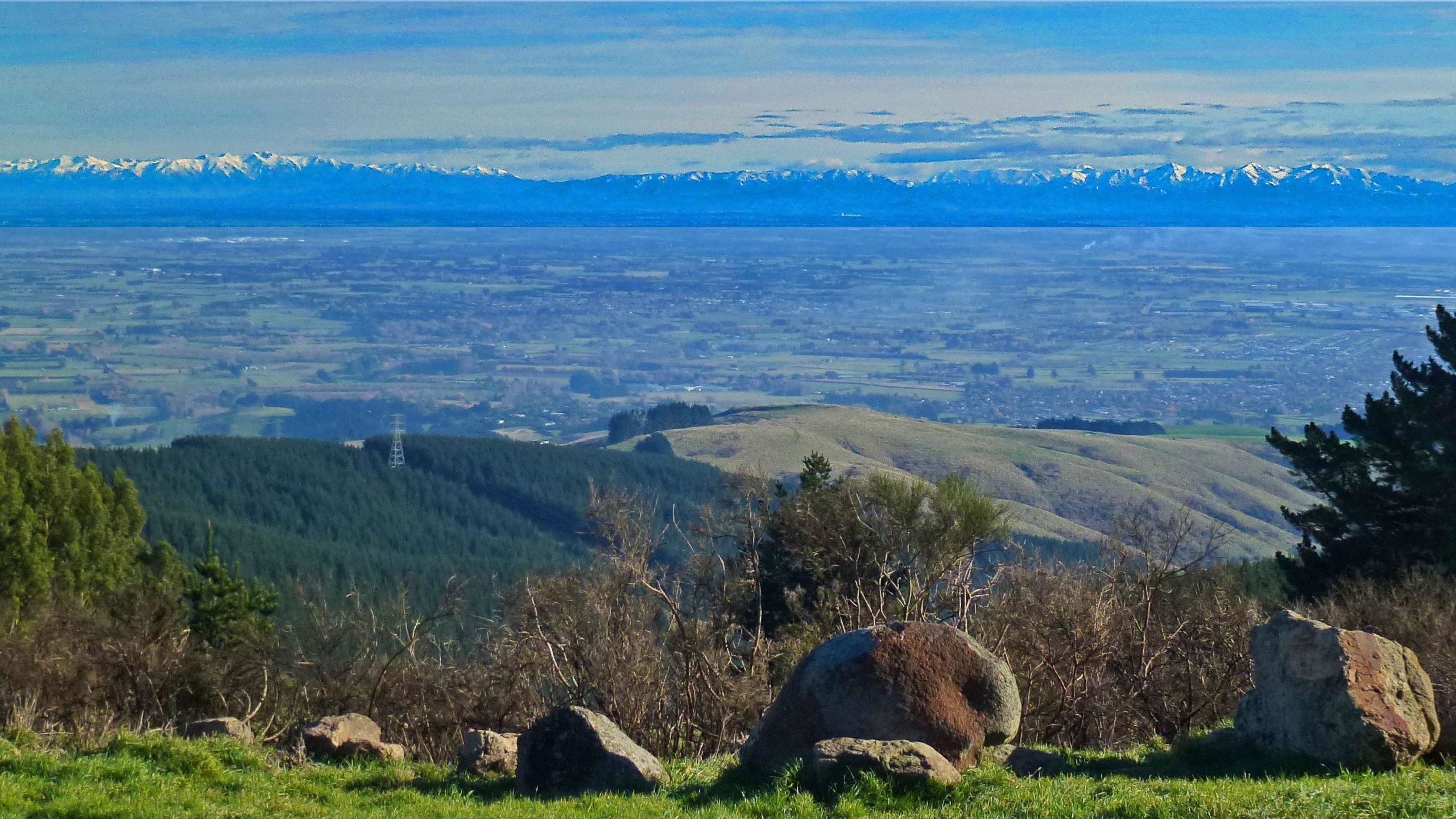 A panoramic view of a lush green valley bordered by rolling hills and fields, with a distant mountain range capped in snow under a clear blue sky. In the foreground, several large rocks are scattered amidst greenery, providing a natural setting that highlights the vast landscape. Port Hills Tracks mountain bike trail.