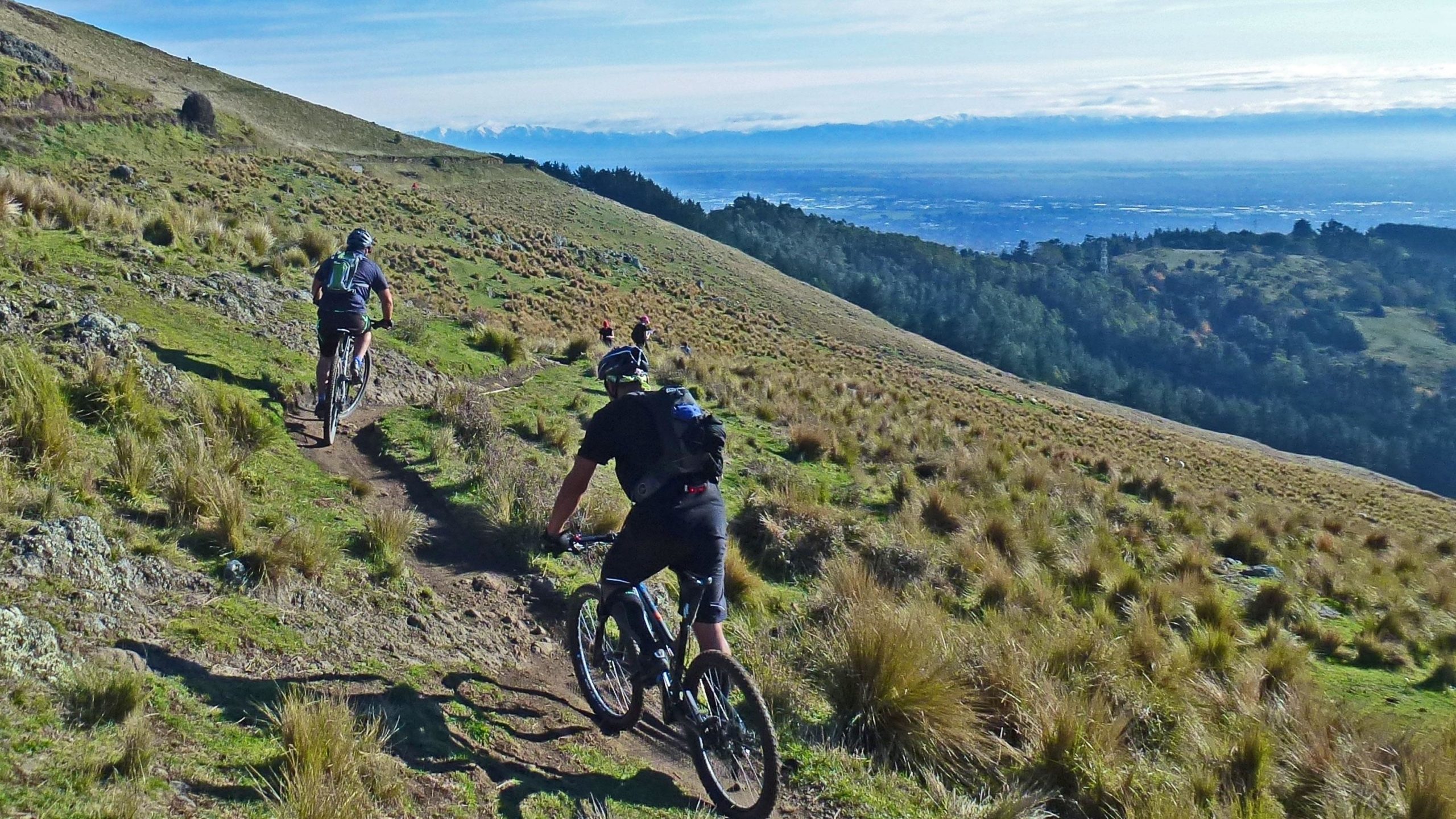 A group of mountain bikers riding along a dirt trail on a hillside, surrounded by grass and rocks. In the background, rolling hills and distant mountains are visible under a clear blue sky. Port Hills Tracks mountain bike trail.
