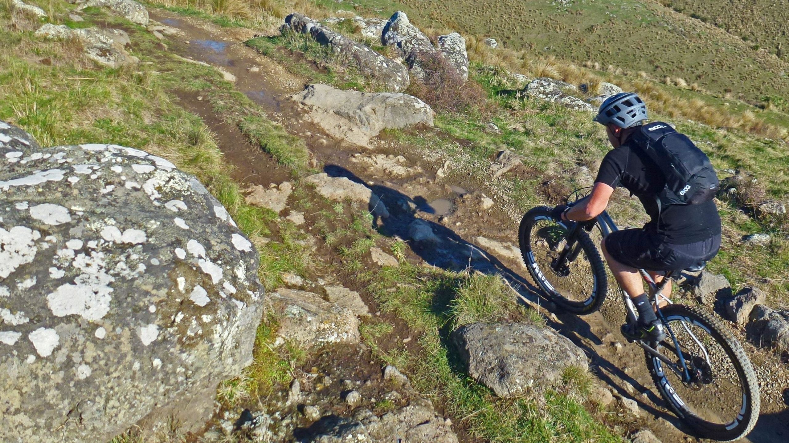 A mountain biker navigating a rocky trail, surrounded by grass and boulders, under clear blue skies. The biker is wearing a helmet and a backpack, demonstrating an adventurous outdoor activity. Port Hills Tracks mountain bike trail.