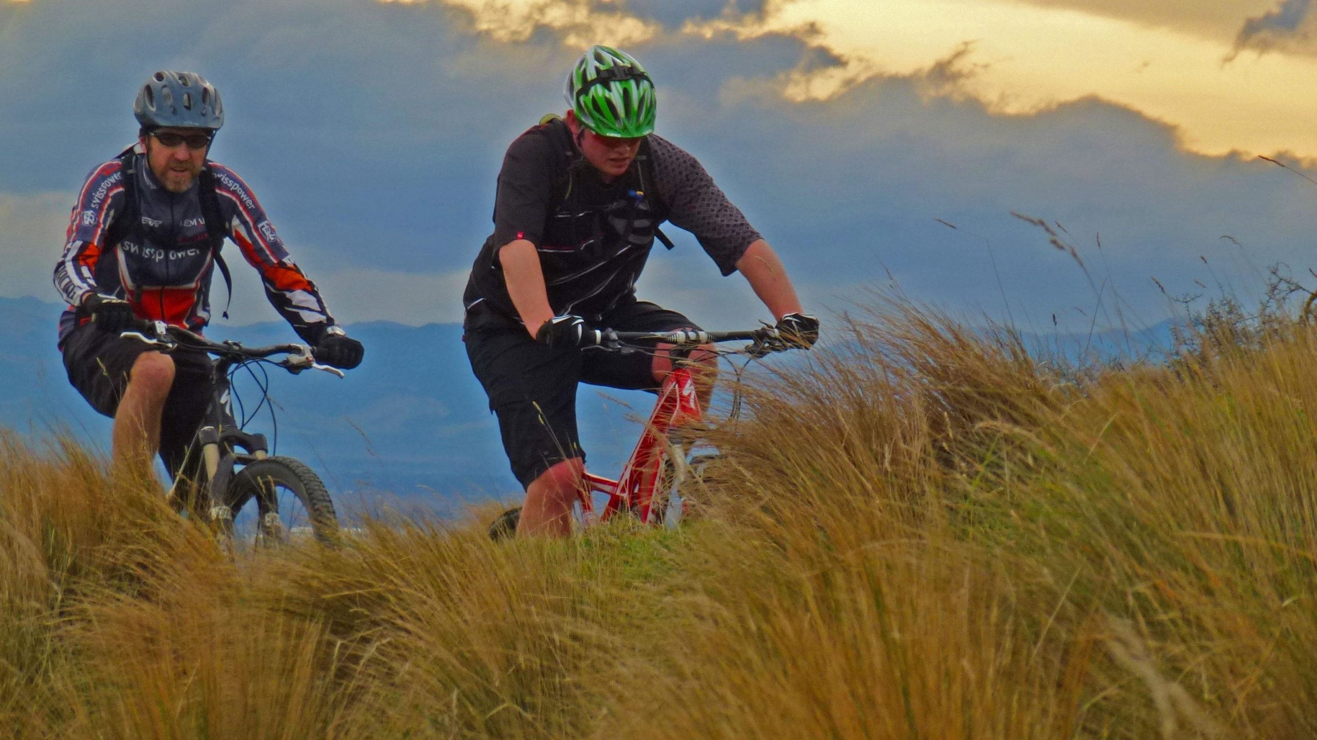 Two mountain bikers navigate a grassy trail, surrounded by tall grass under a cloudy sky. One rider is wearing a helmet and sunglasses with a red and black jersey, while the other is in a green helmet and black gear. The landscape includes distant hills and a dramatic sky. Port Hills Tracks mountain bike trail.