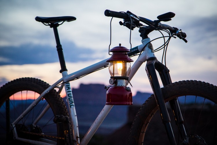 A close-up view of a mountain bike with a vintage lantern hanging from the handlebars, set against a colorful sunset sky in the background. The bike features thick tires and a sturdy frame, while the lantern is lit and adds a warm glow to the scene.