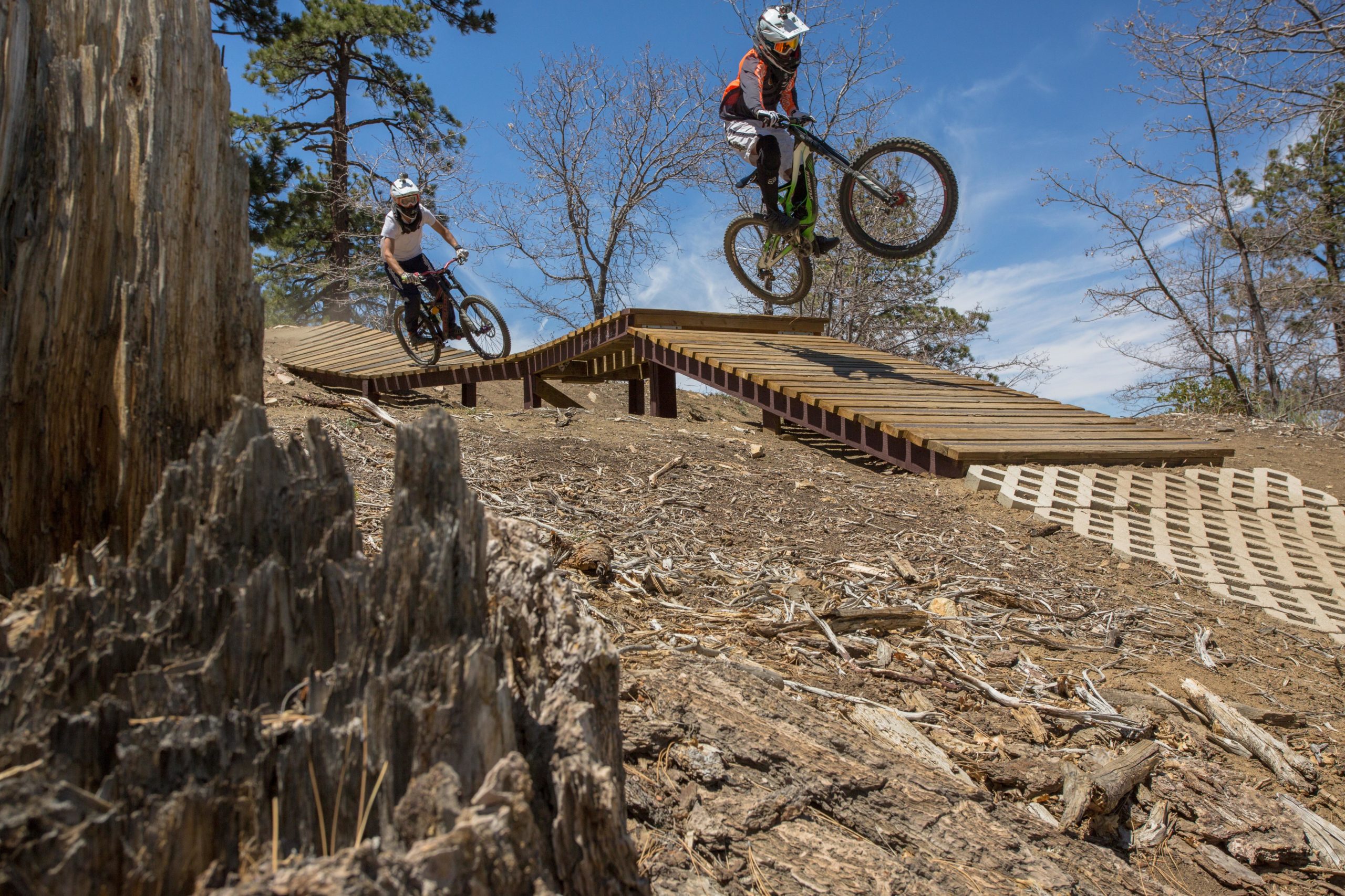 A mountain biker jumps off a wooden ramp on a dirt trail, while another rider descends behind them. The background features trees and a clear blue sky, creating an active outdoor scene. Big Bear Mountain Resort mountain bike trail.