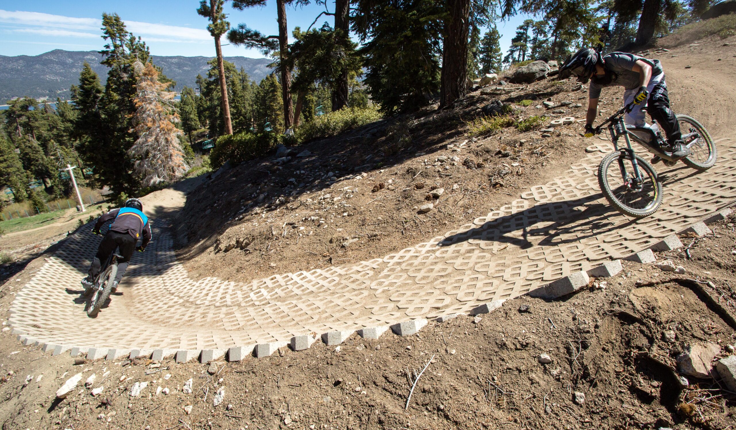 Two mountain bikers navigate a winding dirt trail with a textured surface designed for biking. The scene is set in a hilly, wooded area with trees in the background and mountains visible in the distance under a clear blue sky. Big Bear Mountain Resort mountain bike trail.