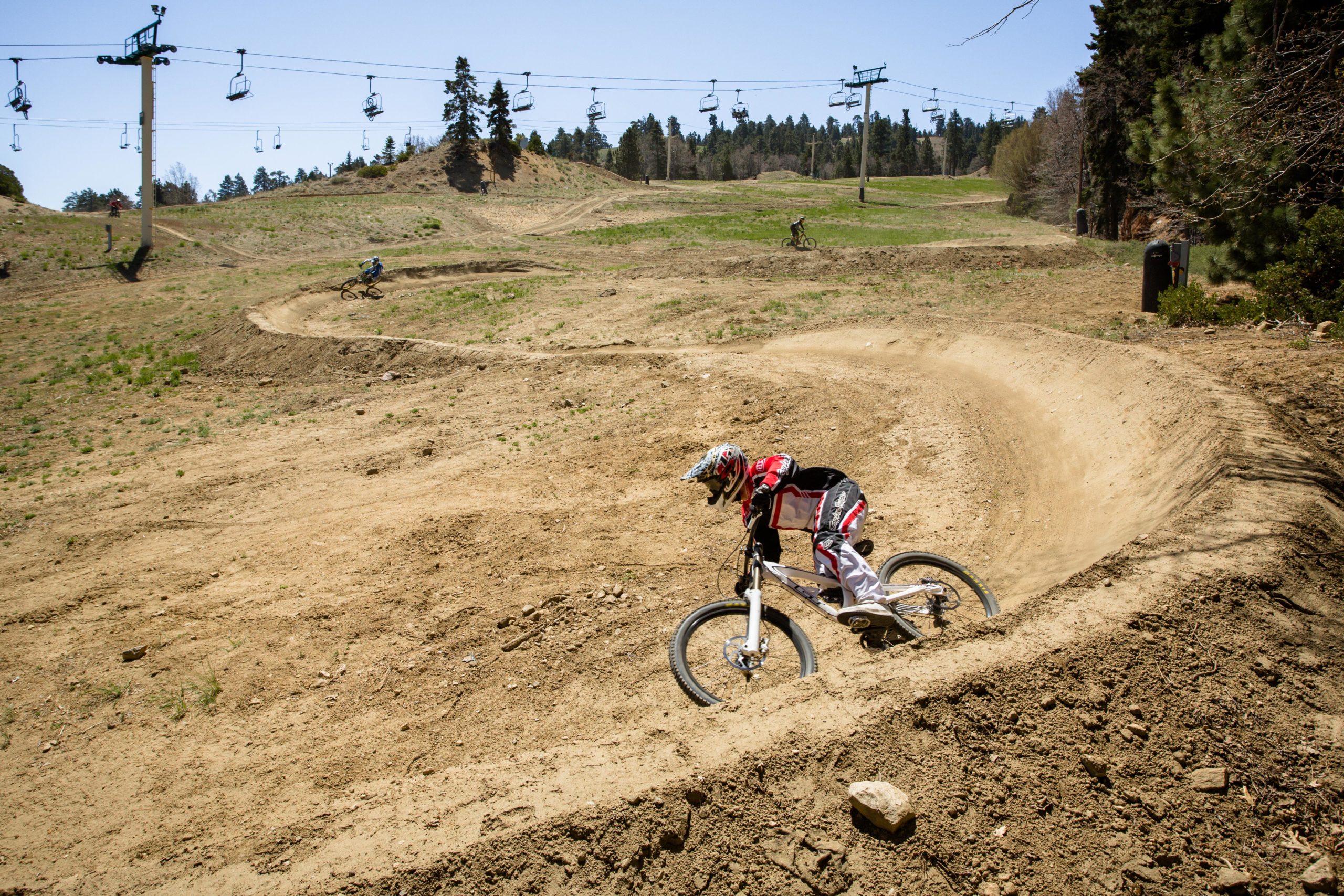 A mountain biker navigates a dirt trail with a turn while other riders can be seen in the background. A ski lift is visible overhead in a clear blue sky, surrounded by trees and grassy areas. Big Bear Mountain Resort mountain bike trail.