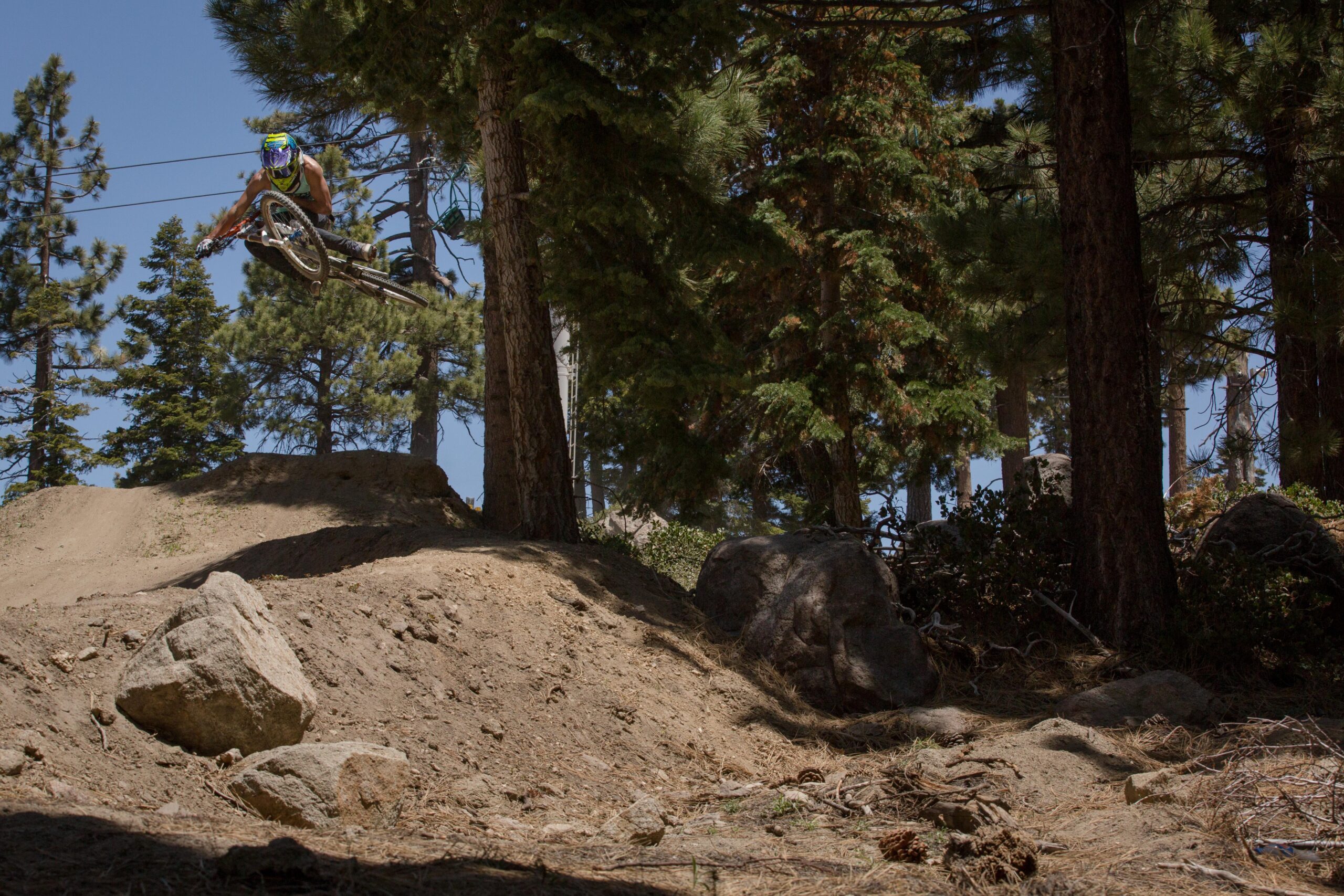 A mountain biker performing a mid-air jump over a dirt ramp, surrounded by tall trees and rocky terrain. The rider is wearing a colorful helmet and gear, showcasing an action shot in a natural outdoor setting. Big Bear Mountain Resort mountain bike trail.