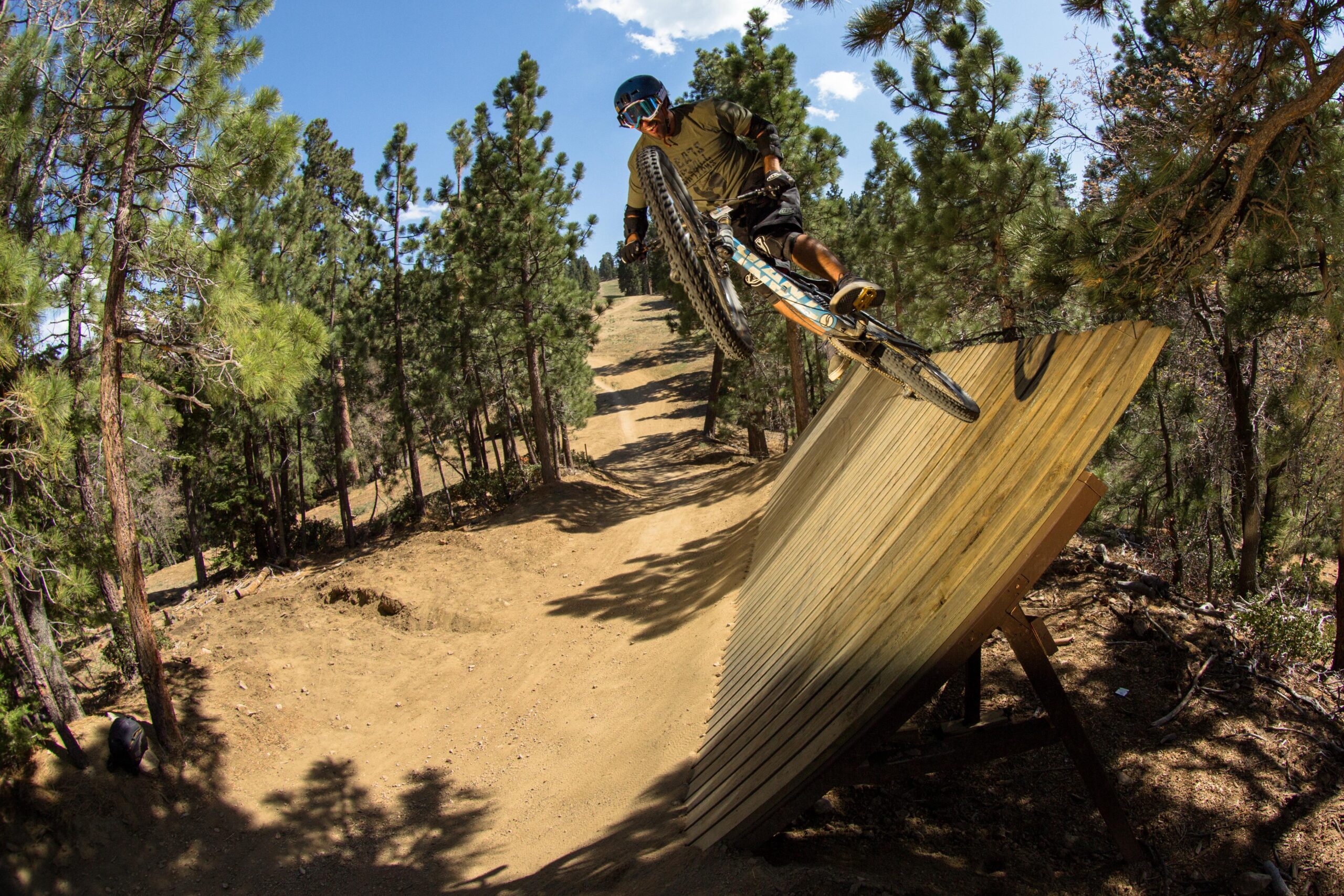 A mountain biker performs a jump off a wooden ramp in a forested area, surrounded by tall trees and a clear blue sky. The rider is airborne, leaning to one side, with their bike wheels elevated above the ramp's edge. Big Bear Mountain Resort mountain bike trail.