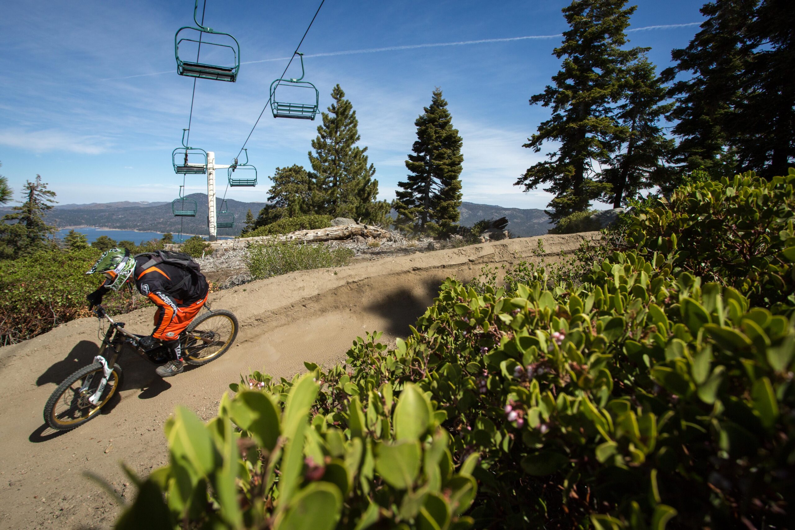 A mountain biker in an orange and black outfit navigates a dirt trail, with a ski lift in the background under a blue sky. Lush green foliage lines the path, and a scenic view of mountains and a lake is visible in the distance. Big Bear Mountain Resort mountain bike trail.