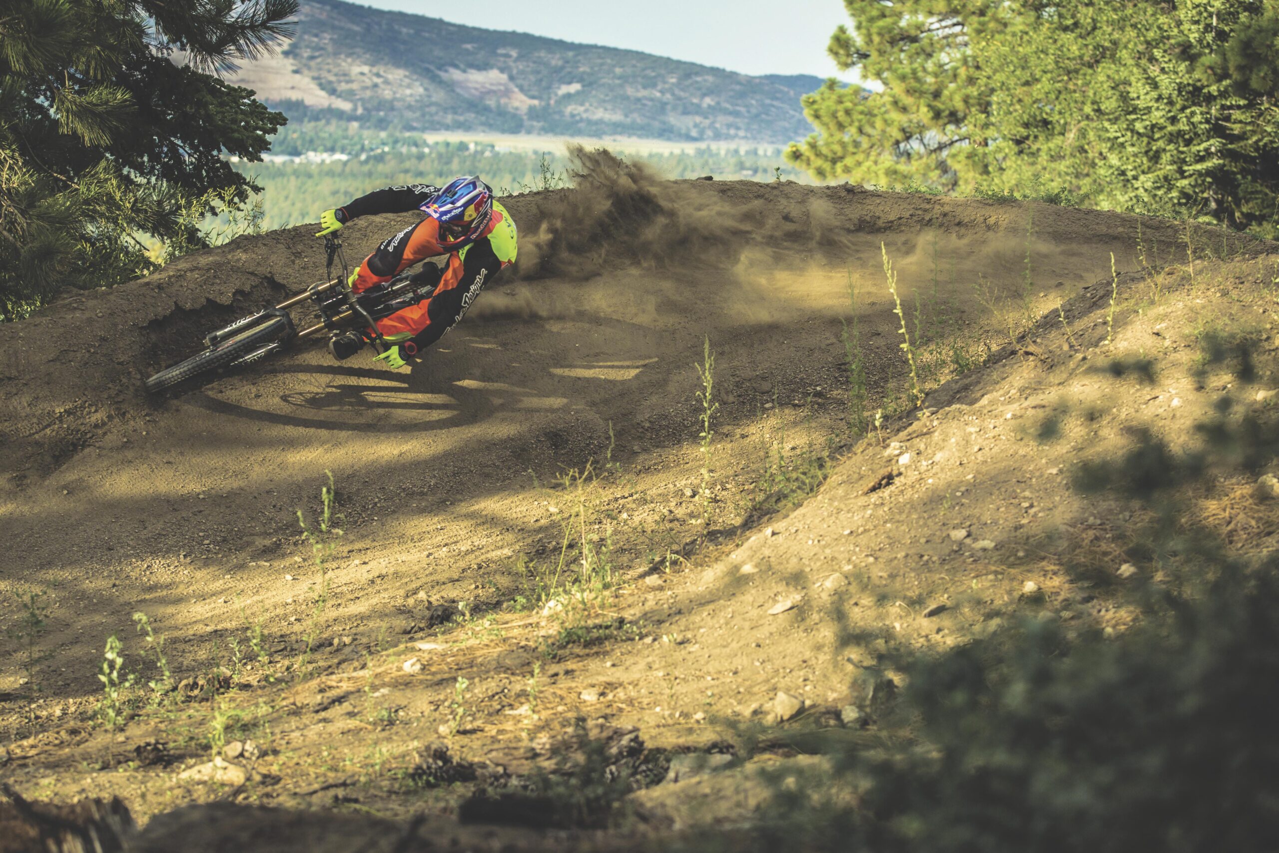 A mountain biker in a colorful outfit leans into a sharp turn on a dirt trail, kicking up dust as he navigates through a forested landscape with hills in the background. Big Bear Mountain Resort mountain bike trail.