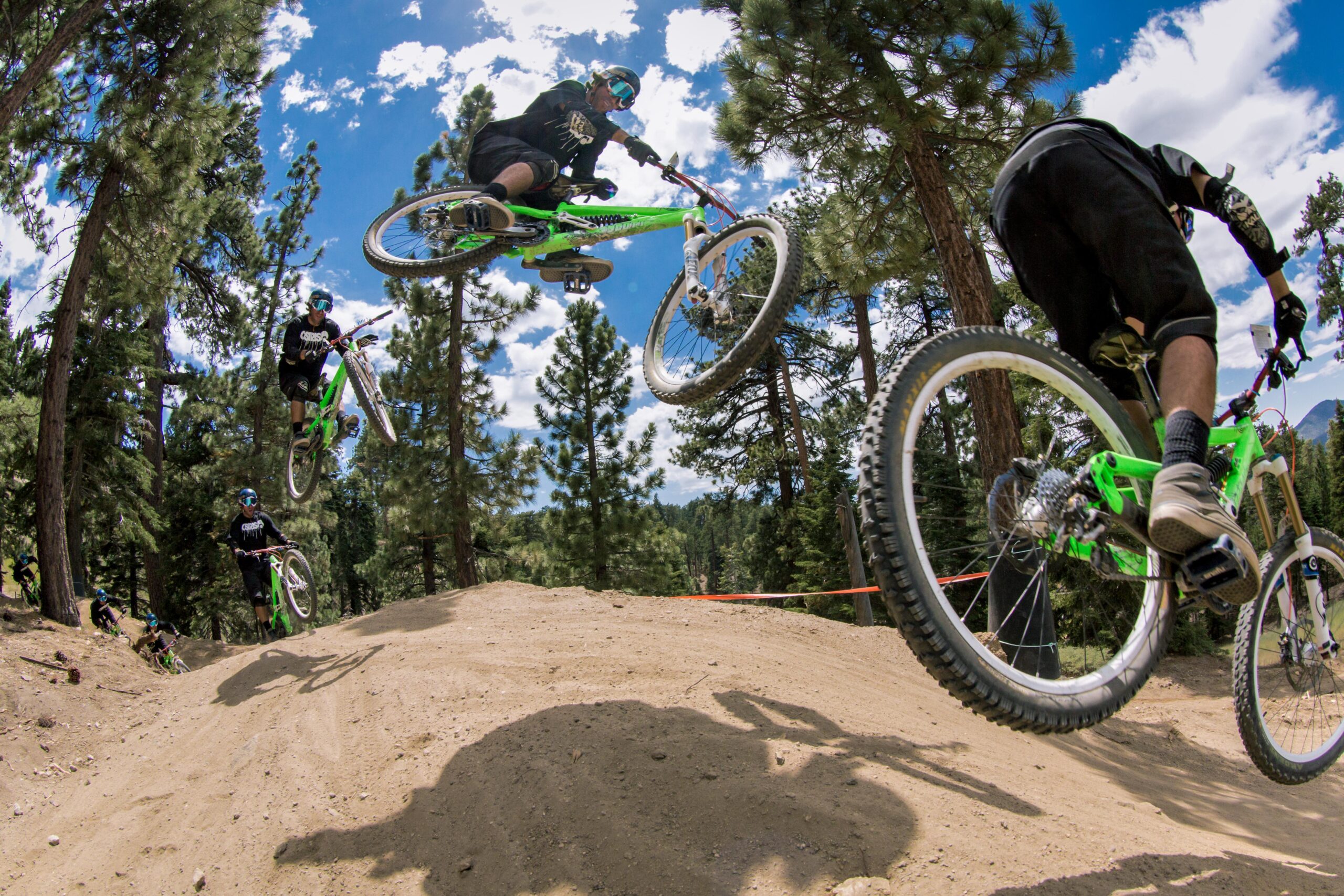 A mountain biker performing a jump over a dirt ramp in a forested area, captured in a sequence showing the rider in mid-air with a blue sky and scattered clouds in the background. The biker is wearing protective gear and riding a green mountain bike. Big Bear Mountain Resort mountain bike trail.