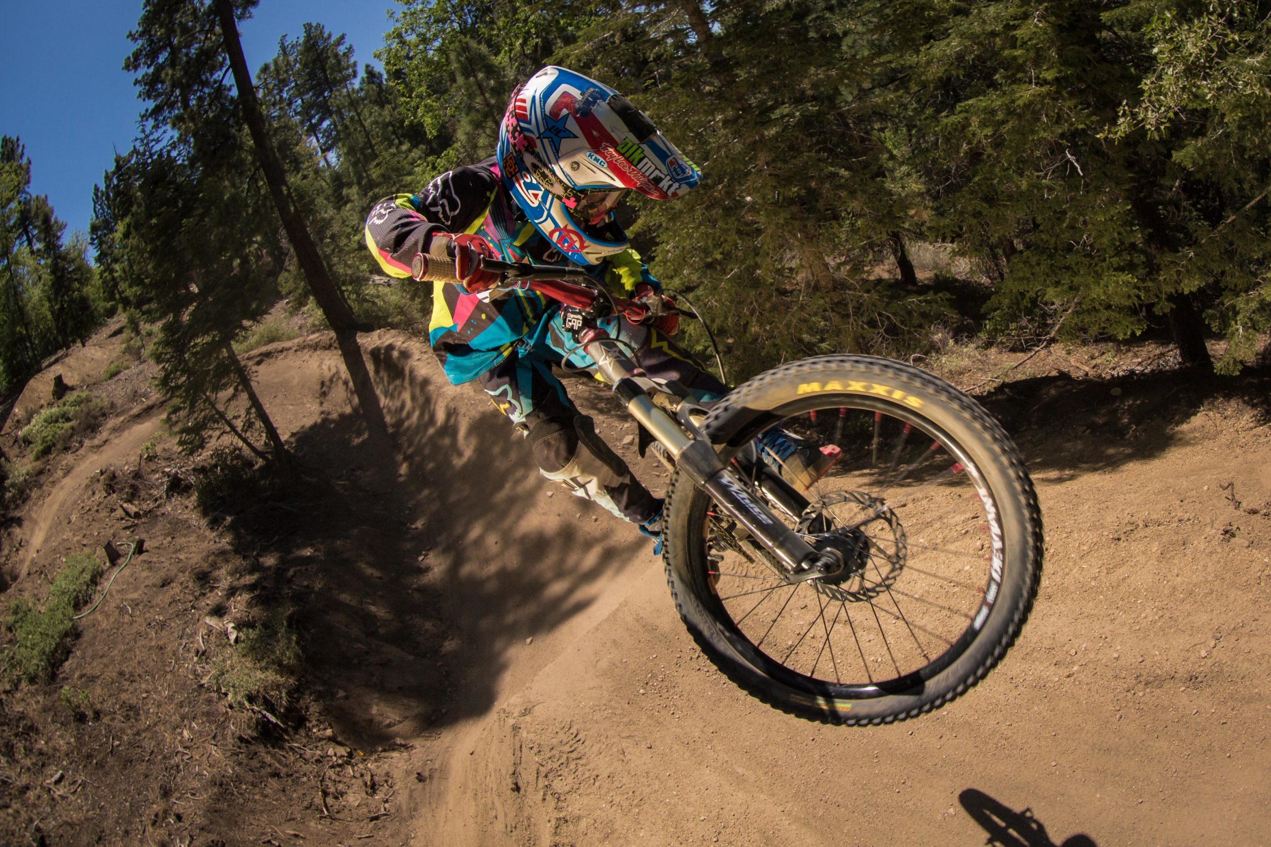 A rider performing a mid-air jump on a mountain bike, showcasing vibrant gear and an action-packed posture against a backdrop of trees and a dirt path. The scene captures the excitement and skill of mountain biking in a natural setting. Big Bear Mountain Resort mountain bike trail.