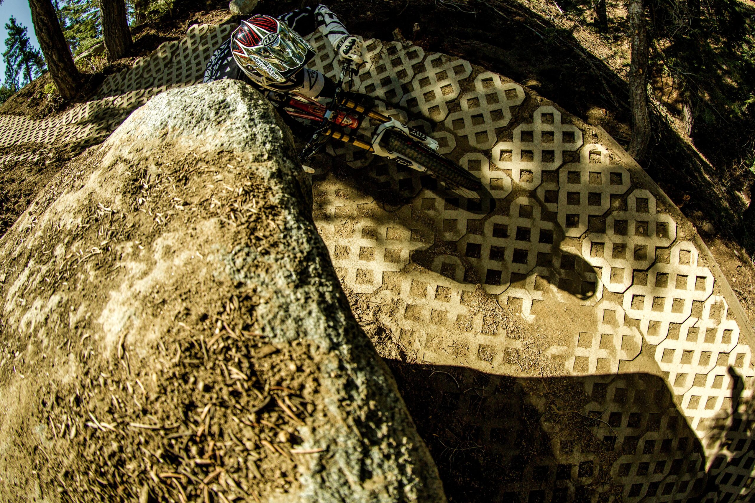 A mountain biker navigating a curved trail, seen from above, with a large rock on the left and a textured, patterned surface on the ground. Trees are visible in the background, creating a natural setting for the biking action. Big Bear Mountain Resort mountain bike trail.