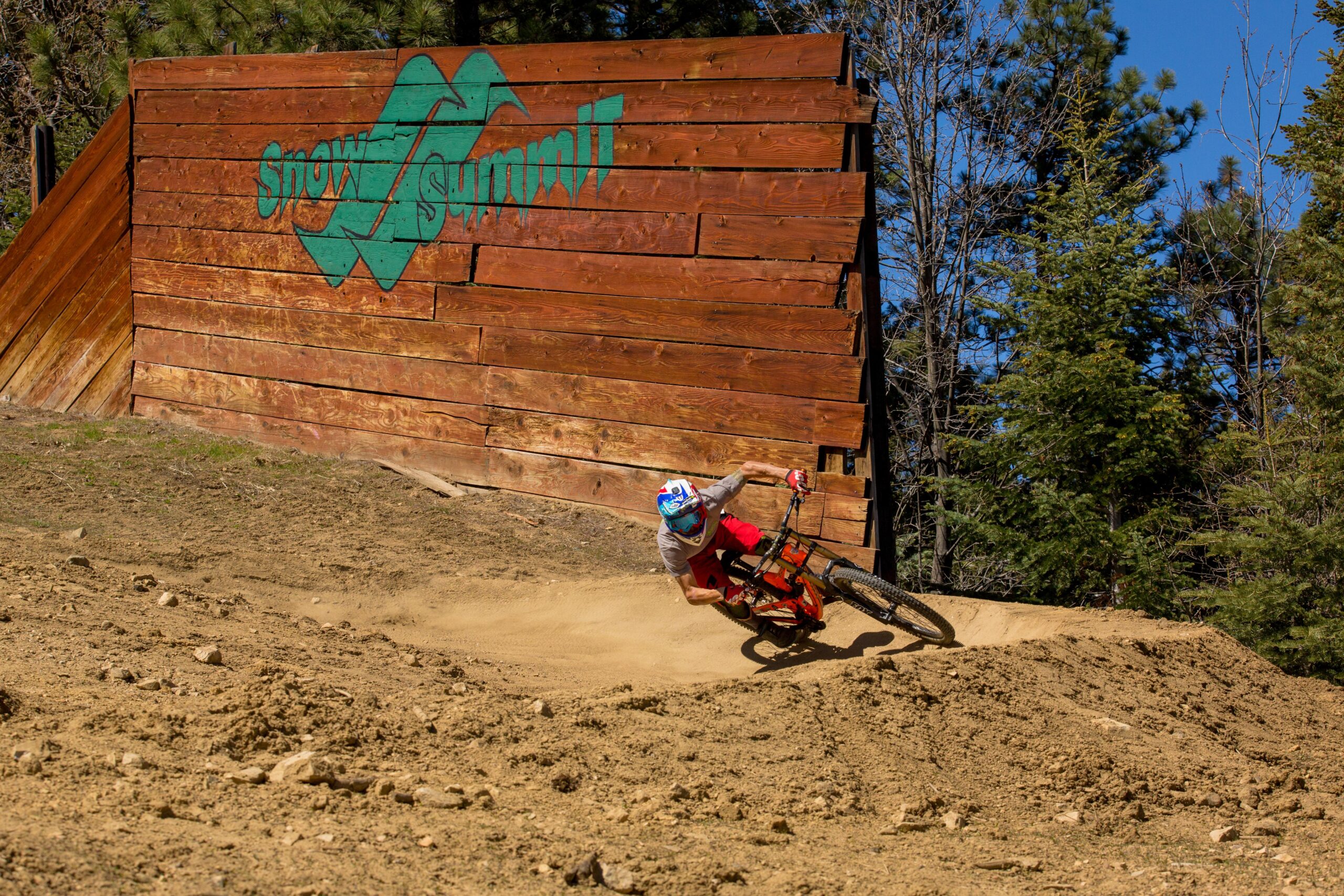 A mountain biker leans into a dirt turn while riding on a trail near a wooden wall with the painted text "Snow Summit." The rider wears a helmet and colorful gear, and the surrounding landscape features trees and a blue sky. Big Bear Mountain Resort mountain bike trail.