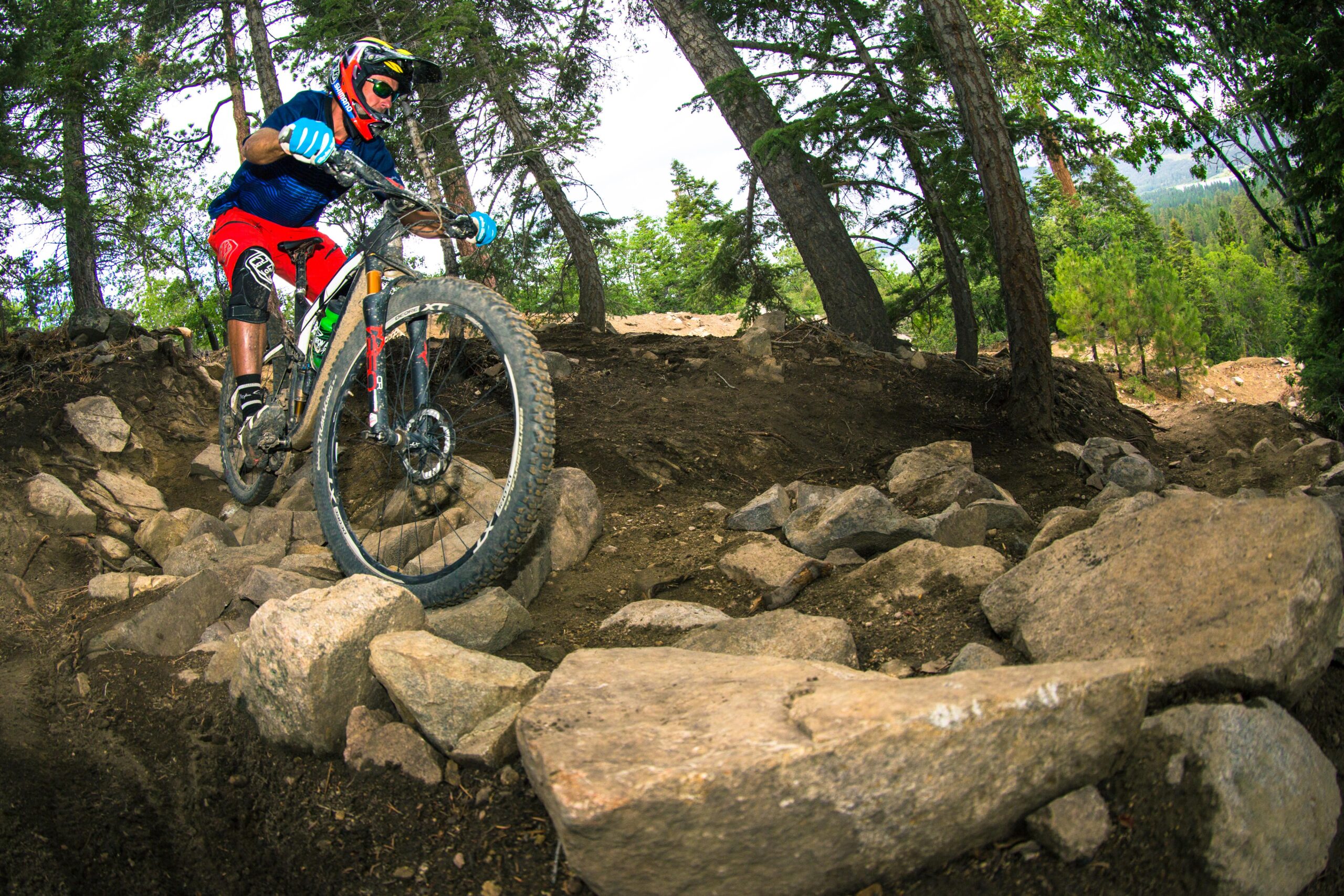 A mountain biker navigating a rocky trail surrounded by trees, demonstrating skill and balance on uneven terrain. The cyclist is wearing protective gear, including a helmet and gloves, and is focused on maneuvering over large stones. Big Bear Mountain Resort mountain bike trail.