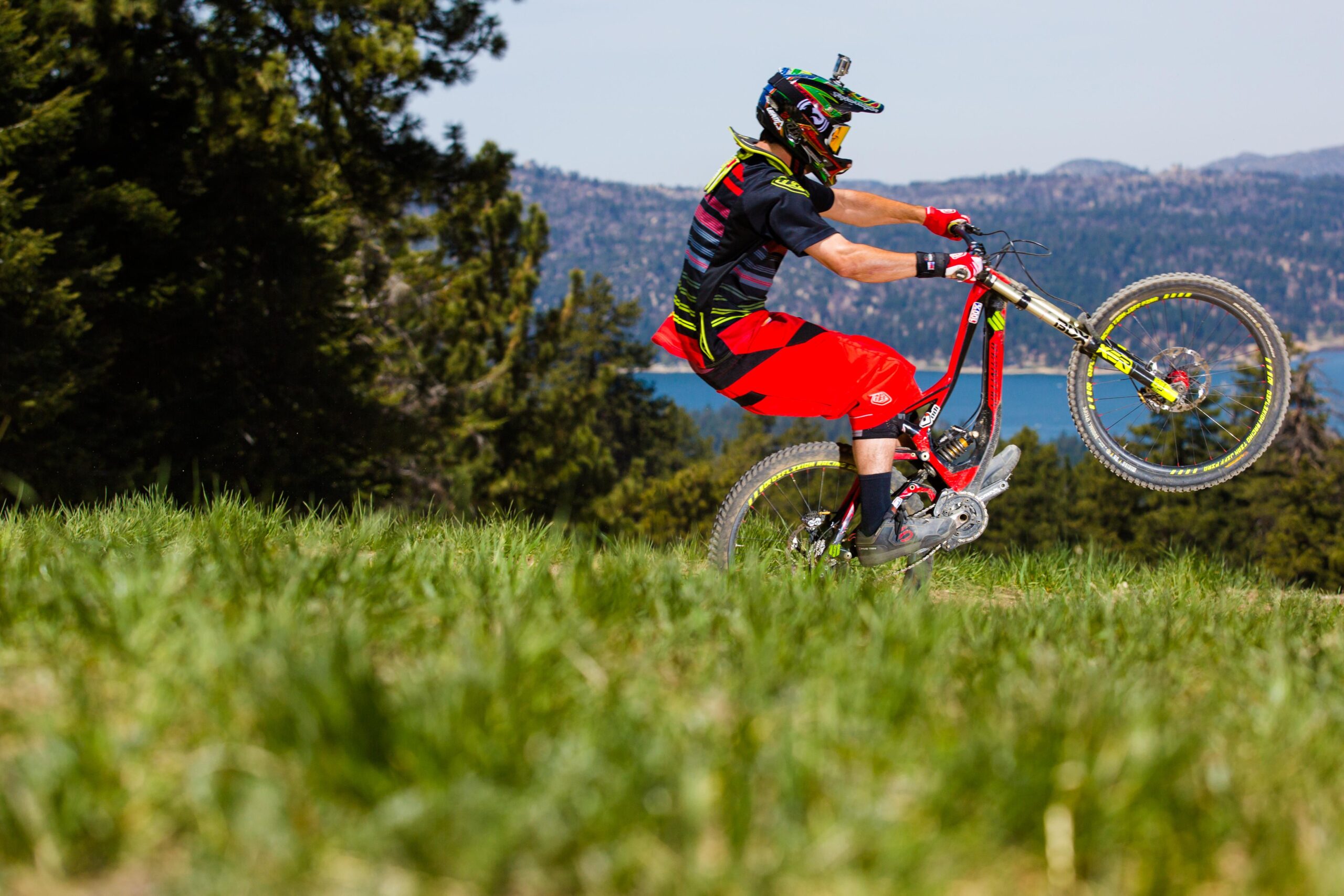 A person doing a wheelie on a mountain bike in a grassy field, surrounded by trees and a scenic mountainous background. The rider is wearing a helmet and protective gear, with a lake visible in the distance under a clear blue sky. Big Bear Mountain Resort mountain bike trail.
