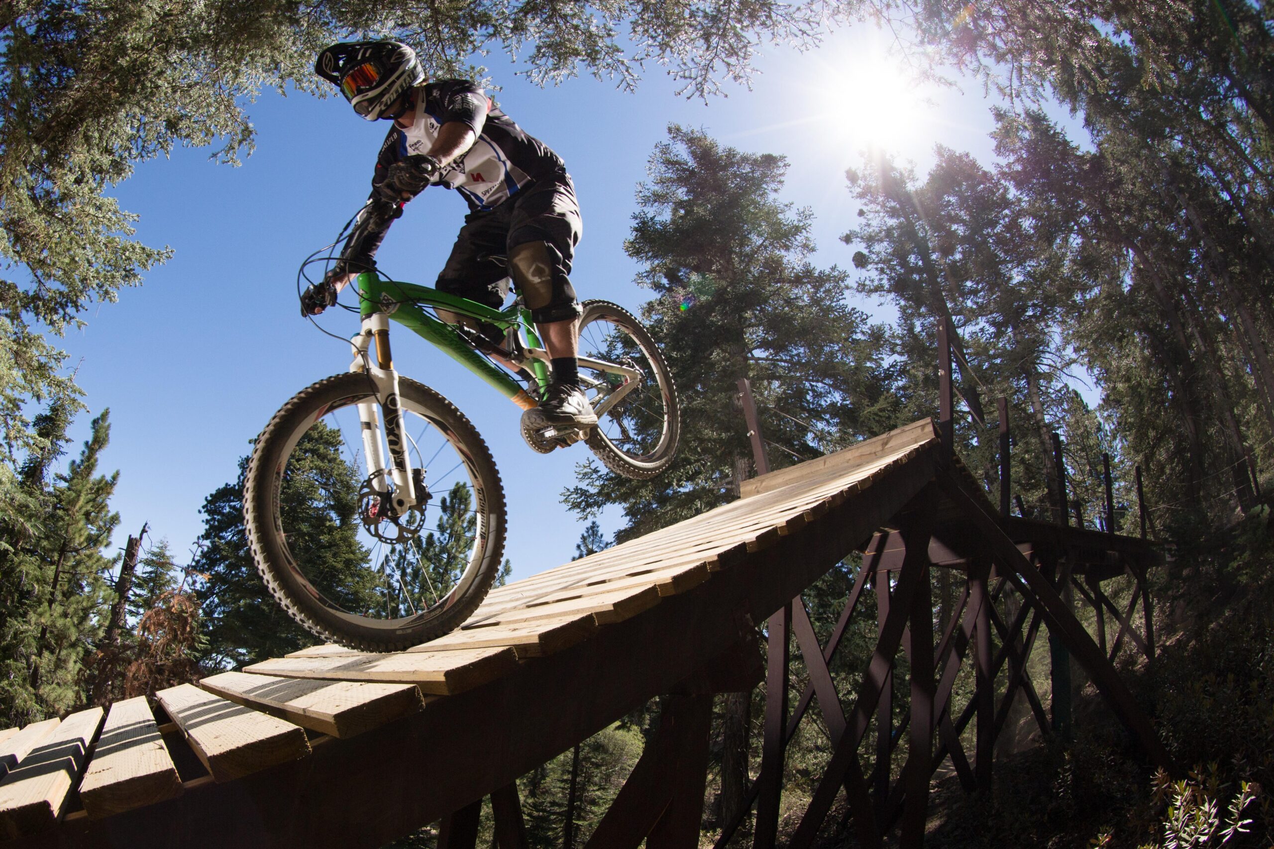 A mountain biker in protective gear is captured mid-air as he jumps off a wooden ramp in a forest setting, surrounded by tall trees and bright sunlight. The bike has a green frame, and the scene conveys a sense of adventure and excitement in the outdoors. Big Bear Mountain Resort mountain bike trail.