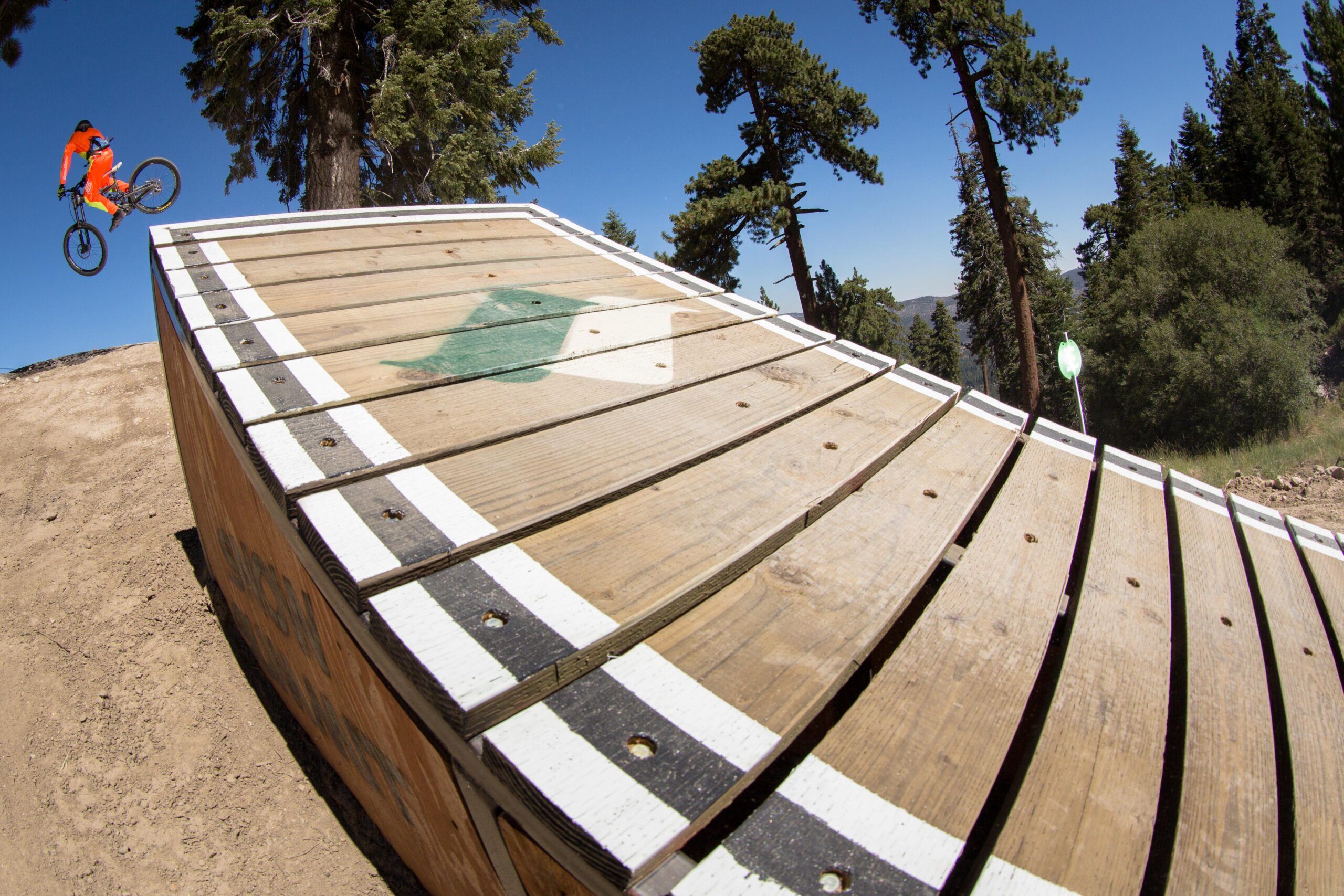 A mountain biker in vibrant orange gear performs a jump off a wooden ramp, surrounded by tall trees under a clear blue sky. The image captures the dynamic moment of the jump with the ramp's details in the foreground, showcasing the thrill of mountain biking. Big Bear Mountain Resort mountain bike trail.