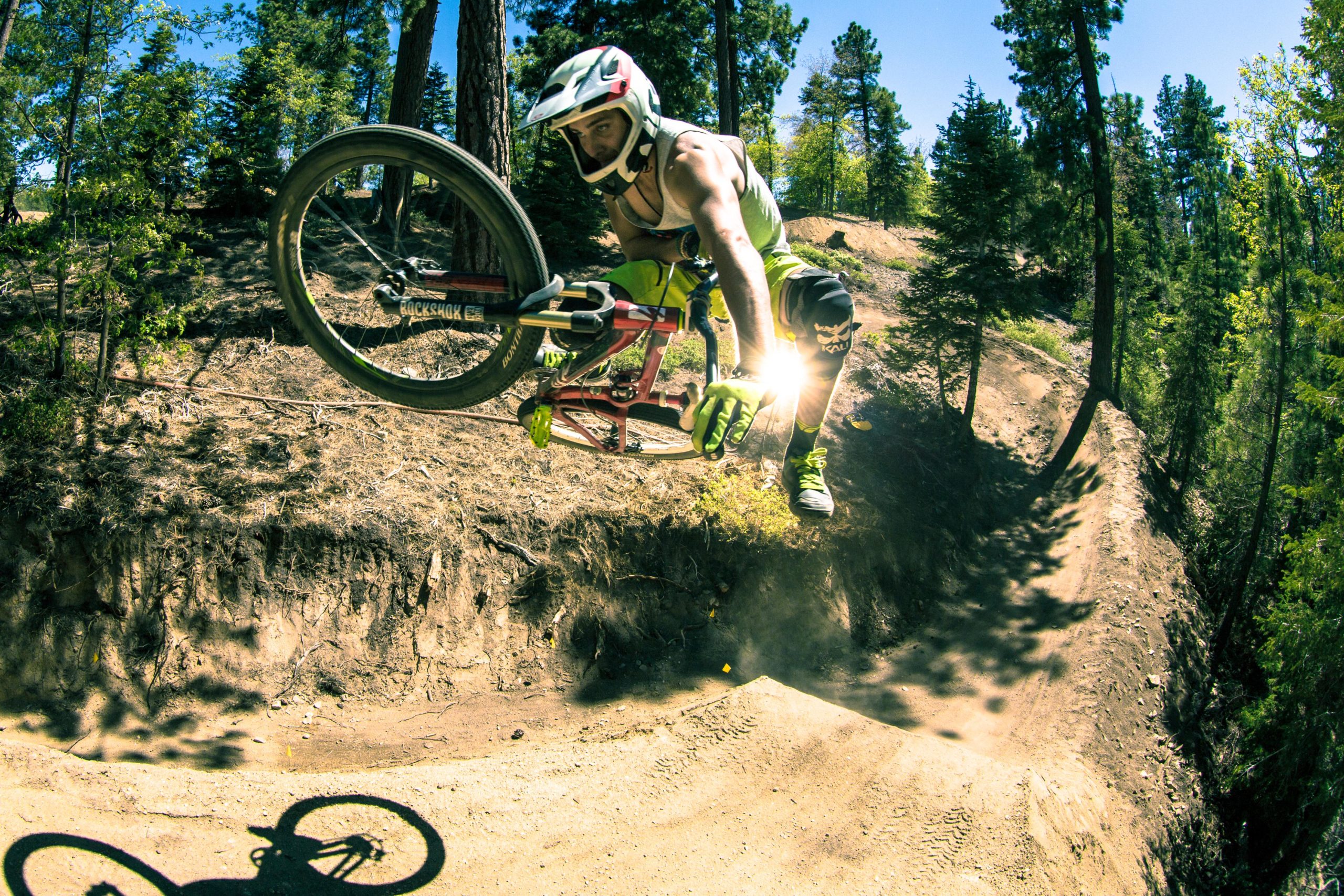 A mountain biker performing a mid-air jump over a dirt ramp in a forested area, wearing a helmet and protective gear, with a shiny sun flare visible near the bike’s tire. Bold trees and bright blue sky are in the background, emphasizing the outdoor setting. Big Bear Mountain Resort mountain bike trail.