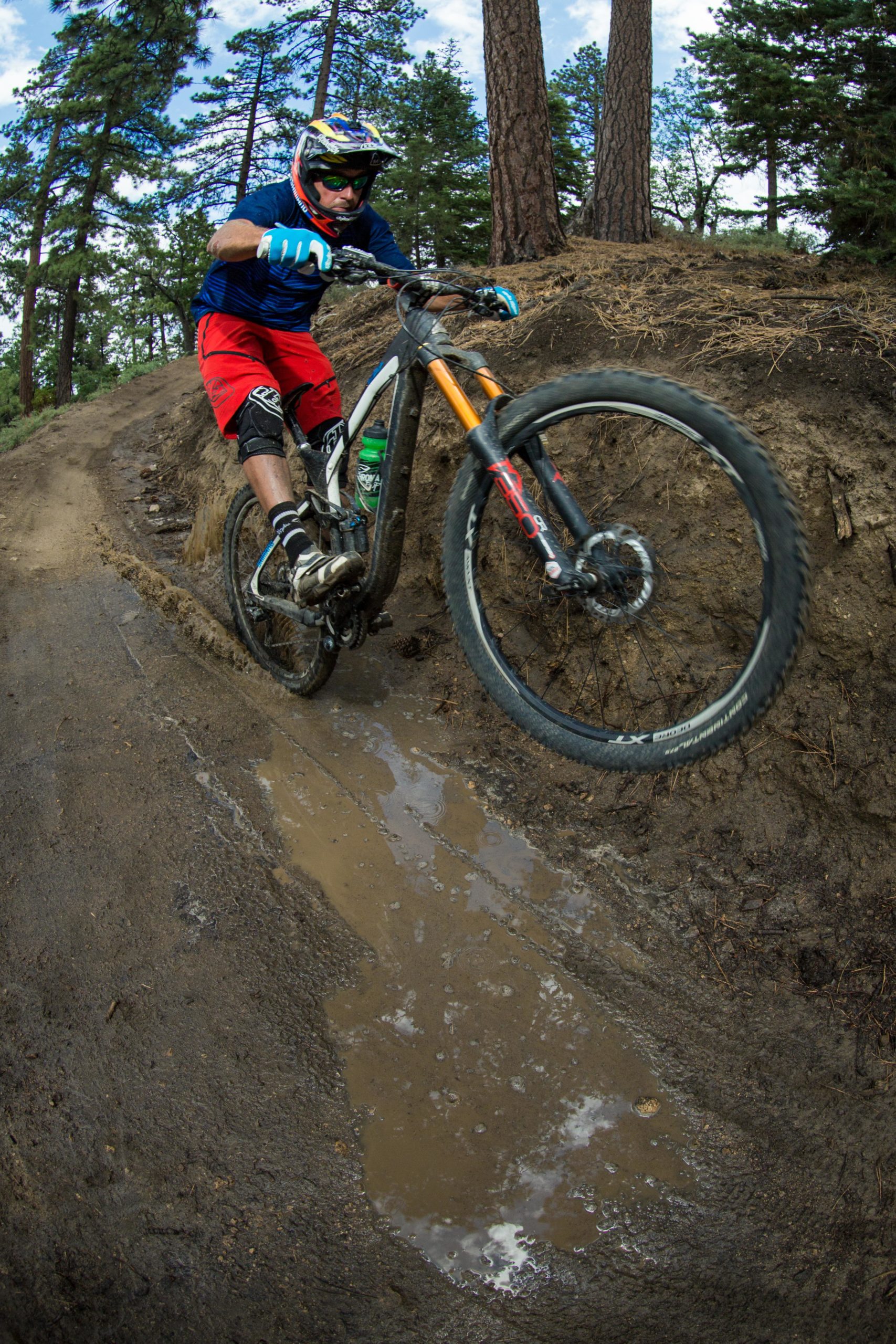 A mountain biker performs a jump over a muddy puddle on a dirt trail surrounded by trees. The rider is wearing a helmet, goggles, and protective gear, showcasing a dynamic and adventurous moment in mountain biking. Big Bear Mountain Resort mountain bike trail.