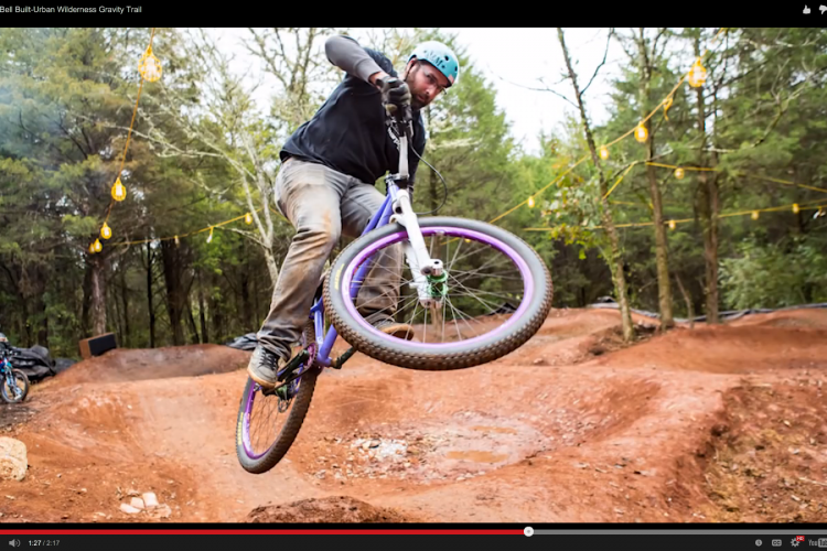 A cyclist performing a jump on a dirt bike trail, surrounded by trees and string lights. The rider is wearing a blue helmet and a black long-sleeve shirt, with a bike featuring purple accents. In the background, another person on a bike is visible, along with a dirt track.