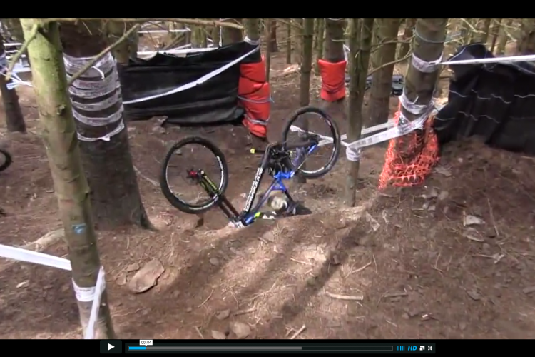 A mountain bike lies on its side in a forested area, surrounded by trees and safety barriers. The ground is covered in dirt and fallen leaves, indicating a biking trail or obstacle course. The bike features a blue frame and is partially hidden among the trees. Safety padding in red and black is placed around some trees, suggesting a downhill biking event.