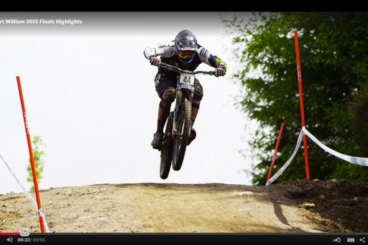 A mountain biker in racing gear performs a jump over a dirt ramp during the Fort William 2015 finals. The biker is airborne with both wheels off the ground, showcasing dynamic movement against a backdrop of trees and a clear sky. Red and white markers are visible on the course.