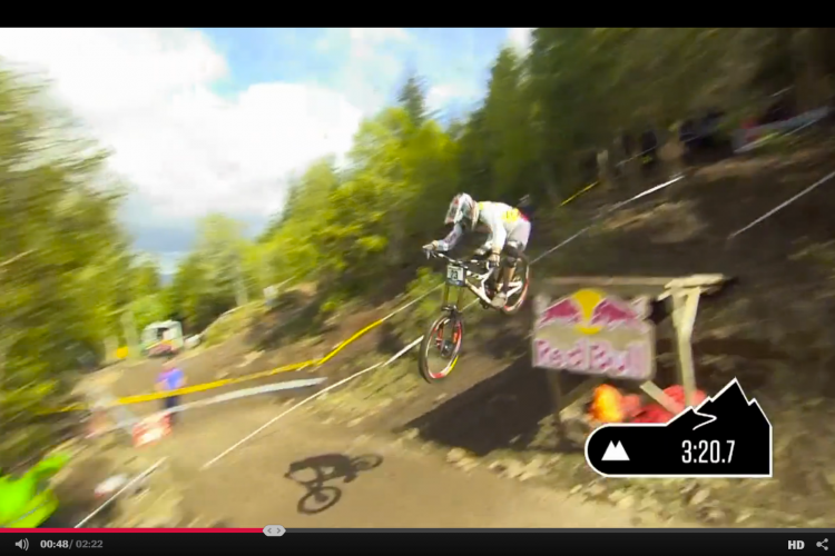A BMX rider performs a mid-air jump on a dirt track, surrounded by trees and spectators. A "Red Bull" sign is visible in the background, and a timer indicating 3:20.7 appears in the corner of the image.